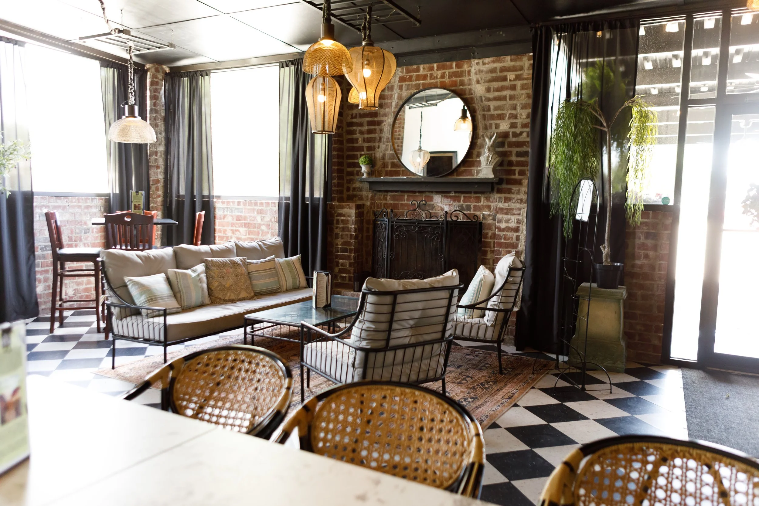 Interior of a cozy café or lounge with brick walls, large windows with black curtains, and eclectic wooden and metal furniture, including a beige couch, rattan chairs, and a small dining table. Hanging pendant lights with woven shades provide warm lighting, and there are decorative plants and a round wall mirror above a fireplace.