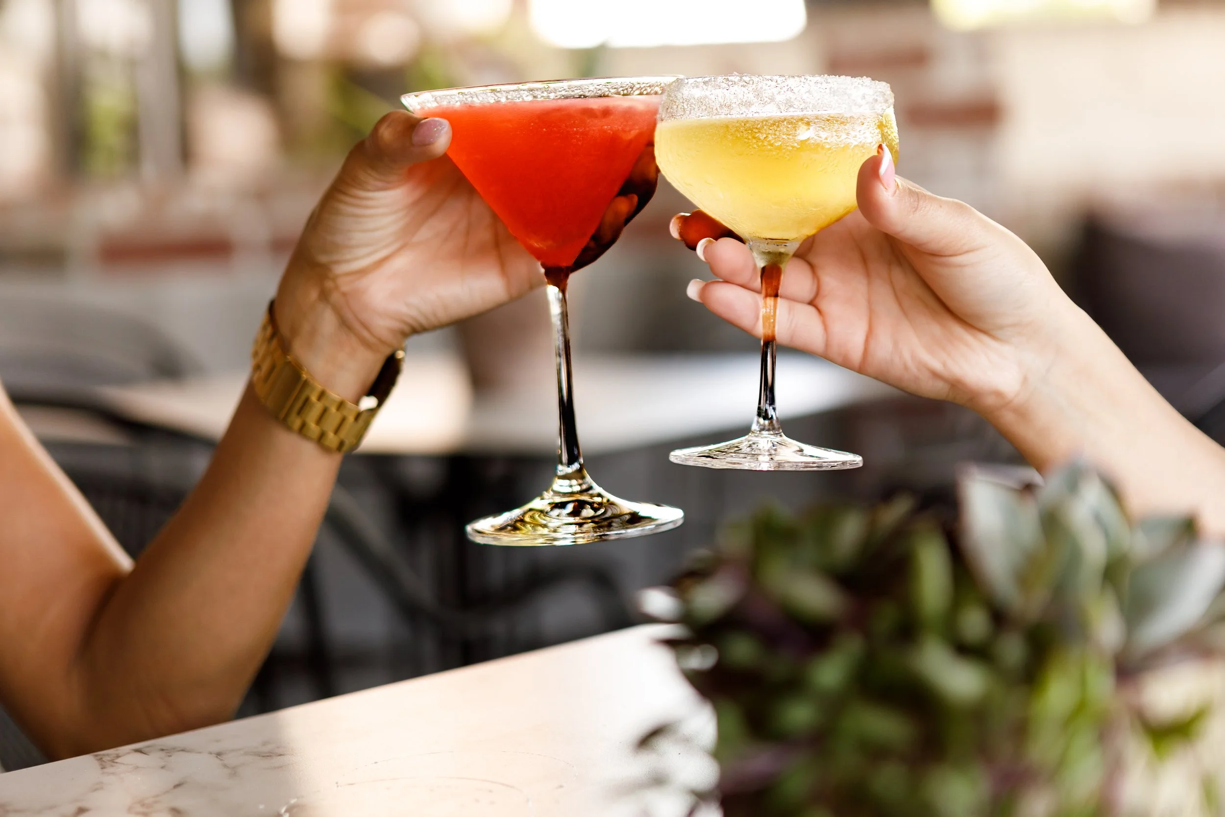 Two hands holding cocktail glasses clinking together. The left hand holds a red drink, and the right hand holds a yellow drink. Both glasses have salted rims, and the scene appears to be indoors with blurred background.