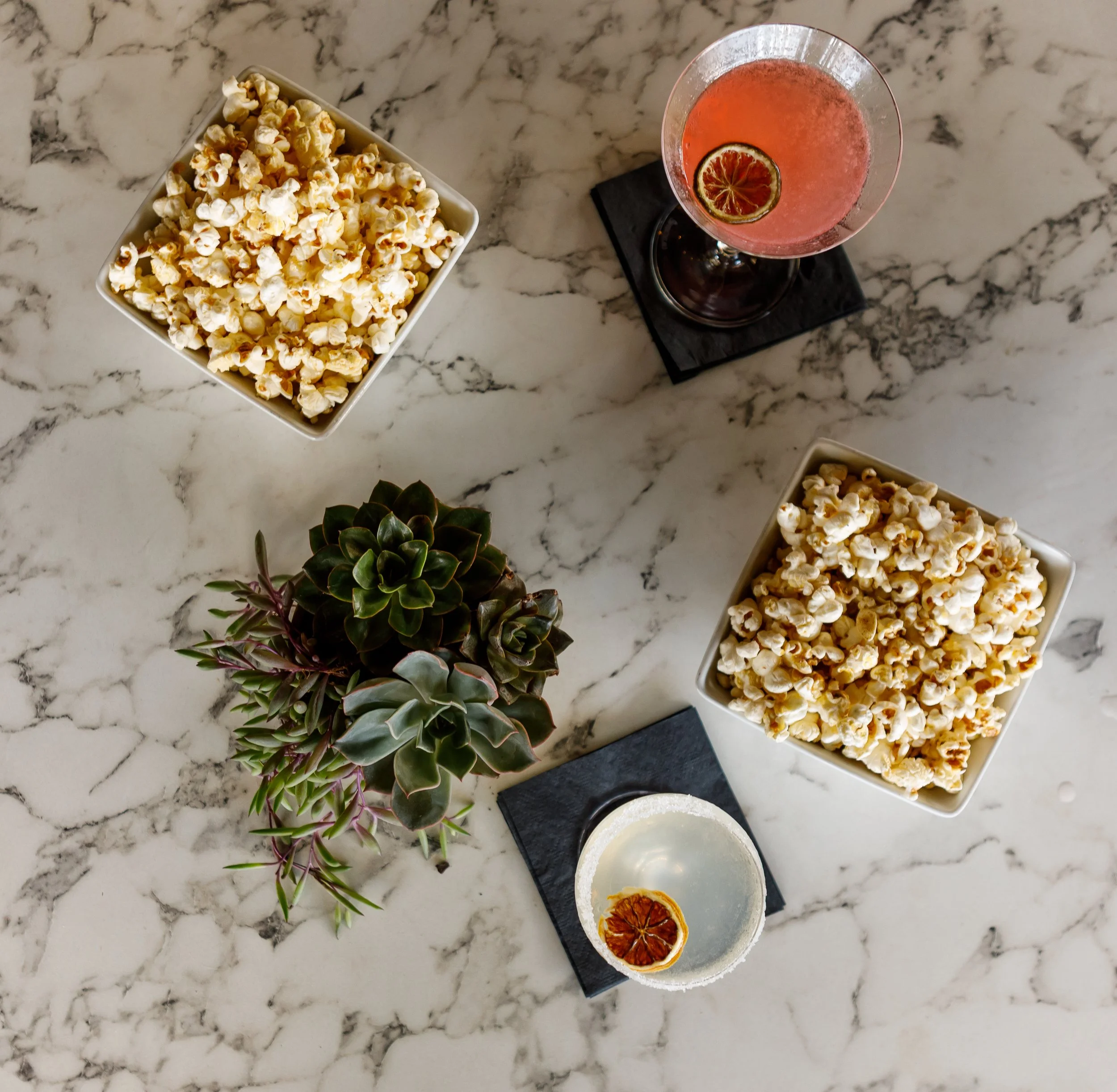 A marble countertop with a bowl of popcorn and a cocktail glass with a pink drink garnished with a dried lemon wheel, along with some succulent plants nearby.