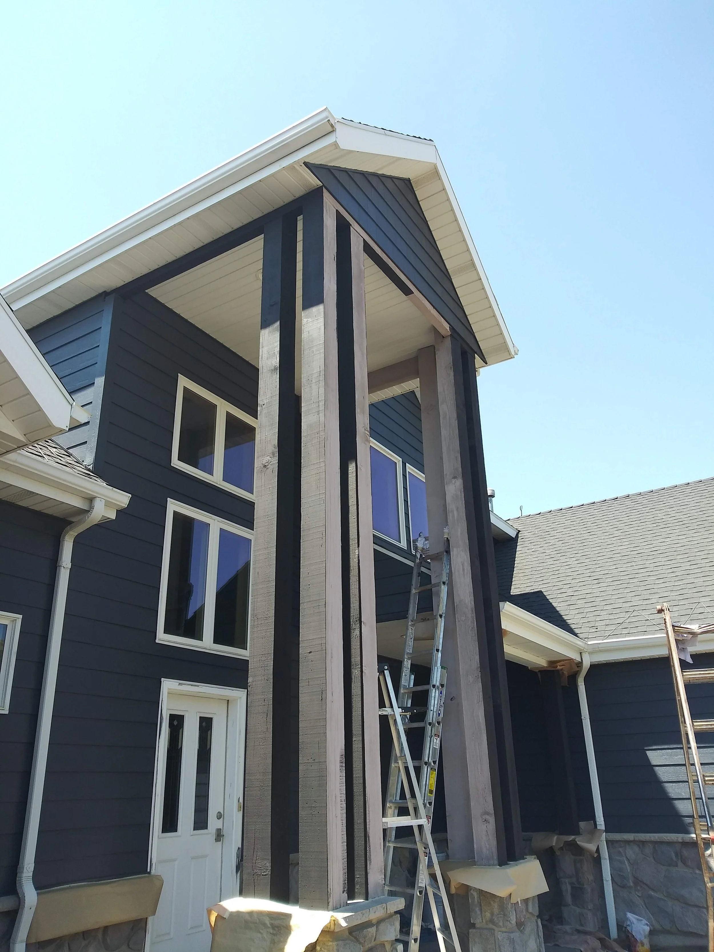Front exterior of a house under construction, showing a new black vertical board siding section with unfinished detailing, ladder, and part of the original house with windows.