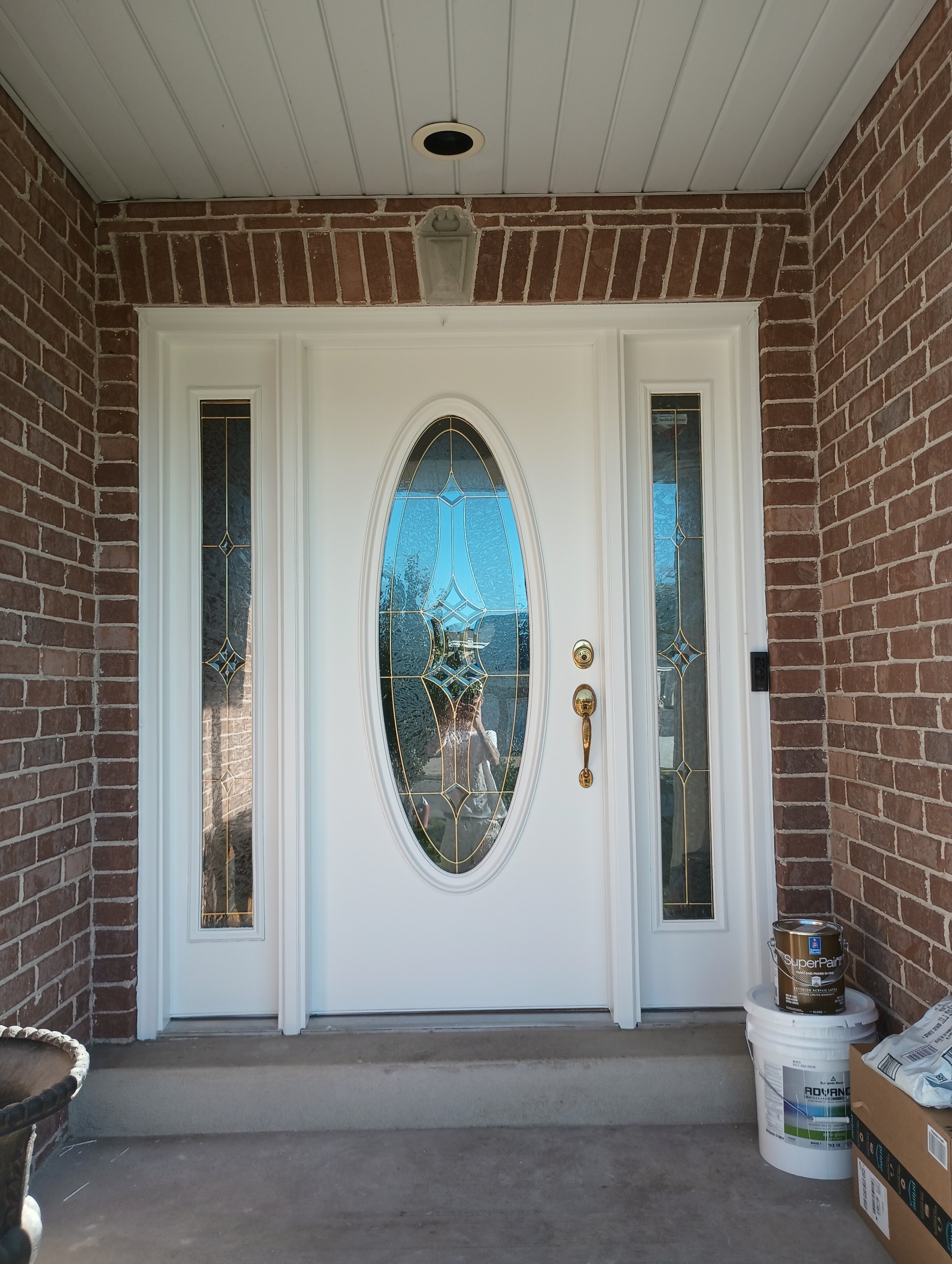 Front door with decorative glass panel, brick walls on sides, white trim, and a porch with some paint supplies and boxes on the right side.