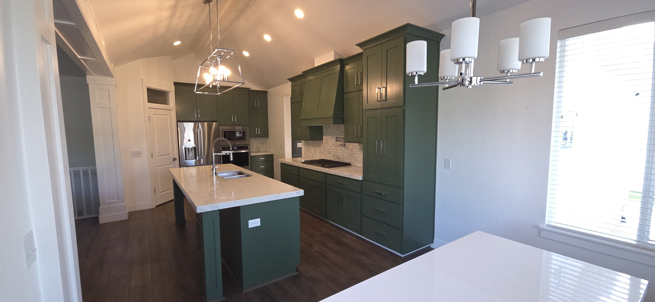 Modern kitchen with green cabinets, white marble countertops, stainless steel appliances, and a large window with blinds