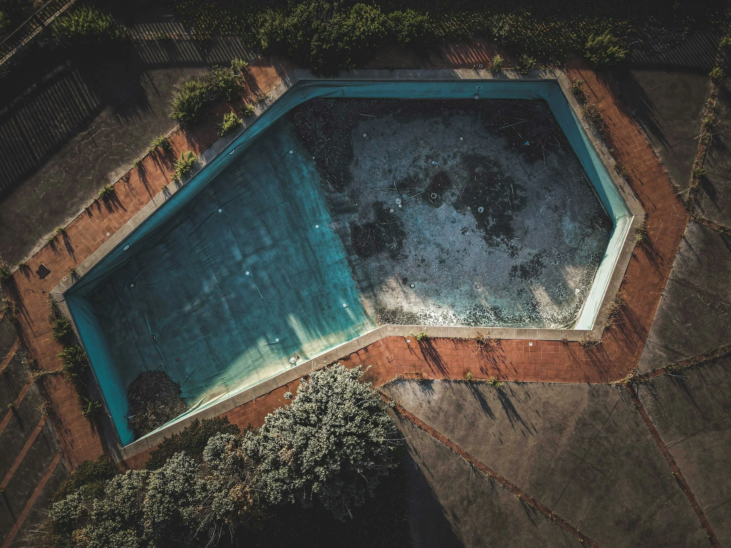 An aerial view of an empty, dilapidated swimming pool with a cracked and stained surface, surrounded by trees and paved walkways.