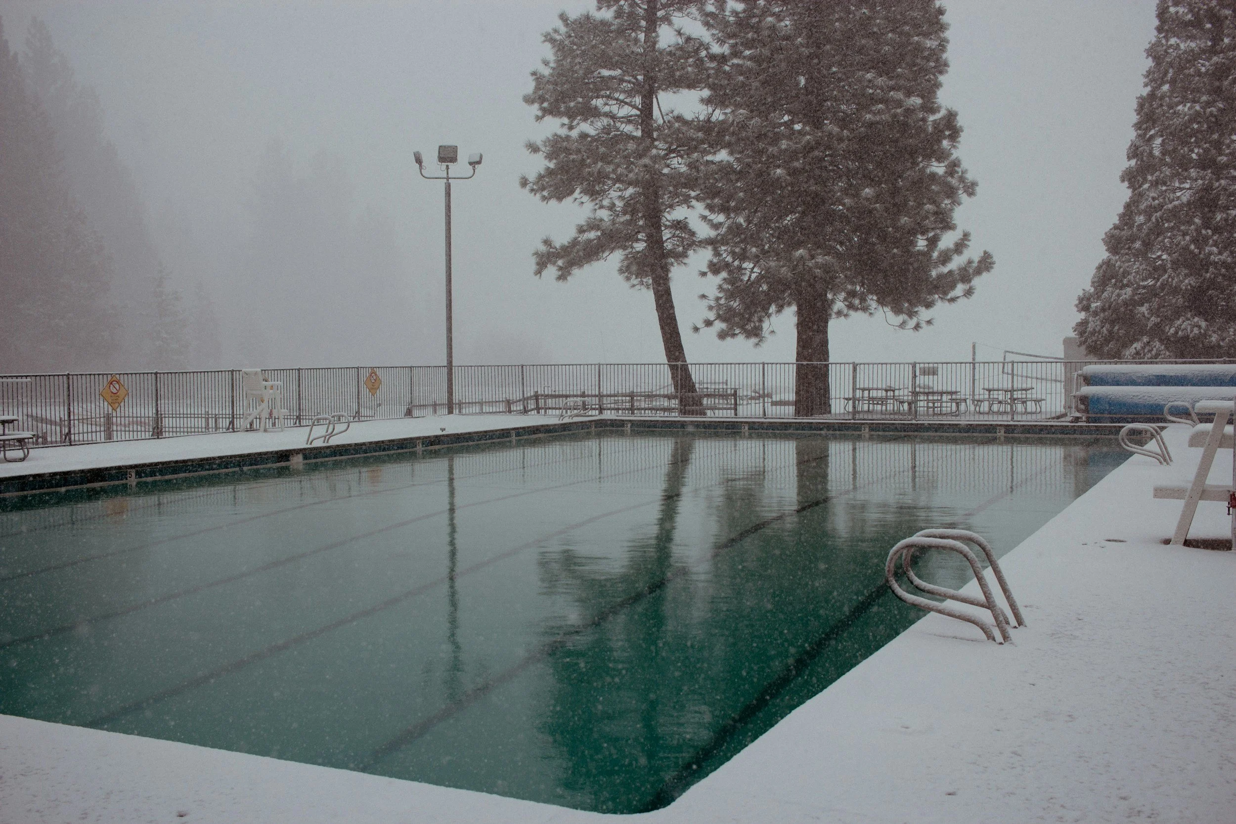 An outdoor swimming pool area during a snowstorm with snow covering the ground and benches. Several trees are visible, covered with snow, and a tall light pole with three floodlights stands near the pool. The pool is empty and the water appears still, reflecting the overcast sky. The scene seems to be cold and quiet, indicating winter weather.