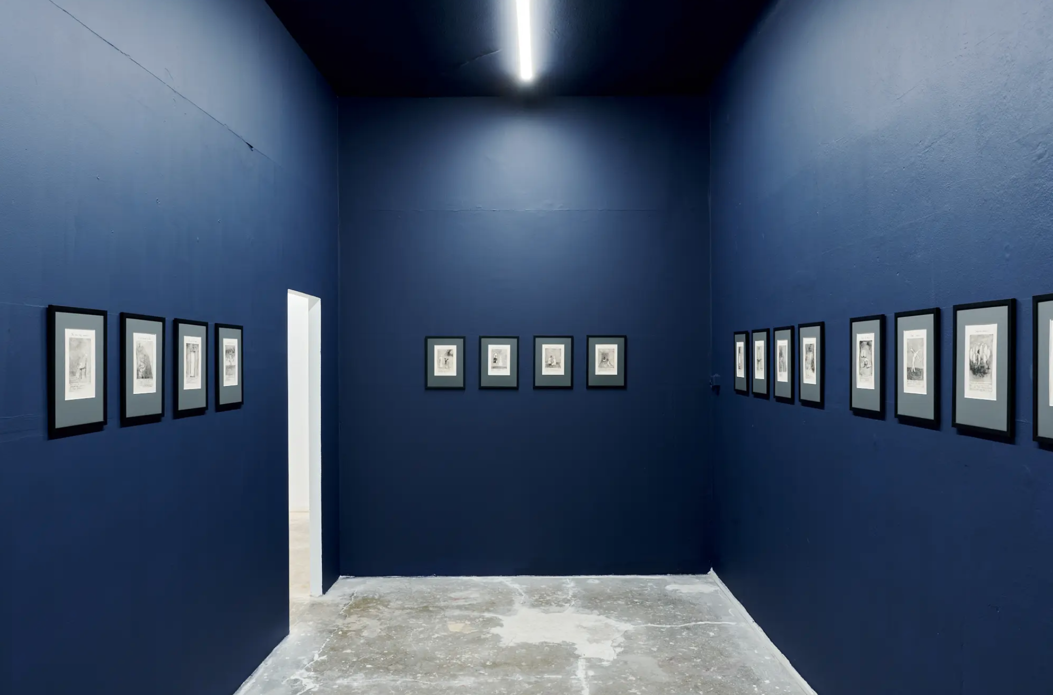 An art gallery with dark blue walls featuring framed black and white artworks on both sides of the corridor, with a doorway visible at the end.