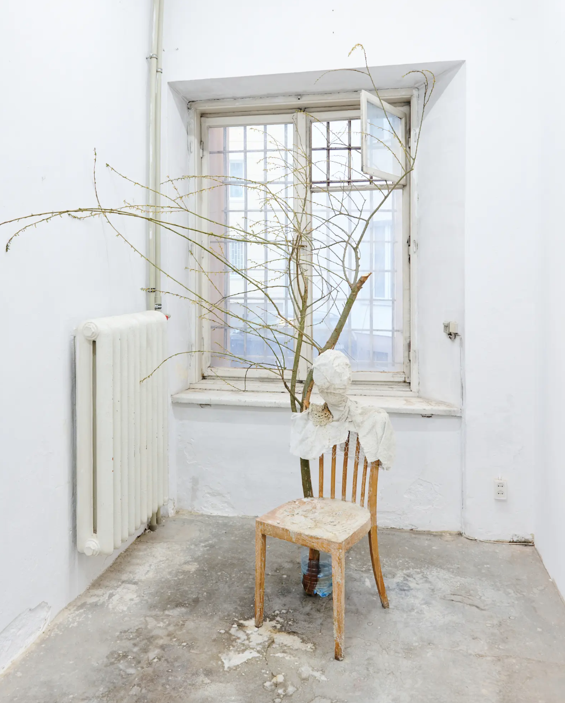 An art installation featuring a wooden chair, a sculpture of a head wrapped in white fabric, and a large, leafless tree branch, set inside a white-walled room with an open window.