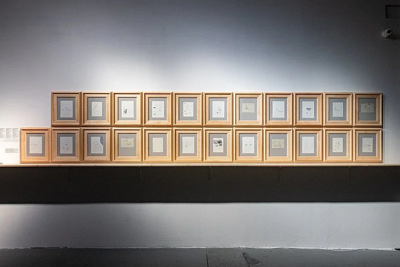 A gallery wall with multiple framed sketches or drawings arranged in a grid, displayed on a white wall with a shelf below.