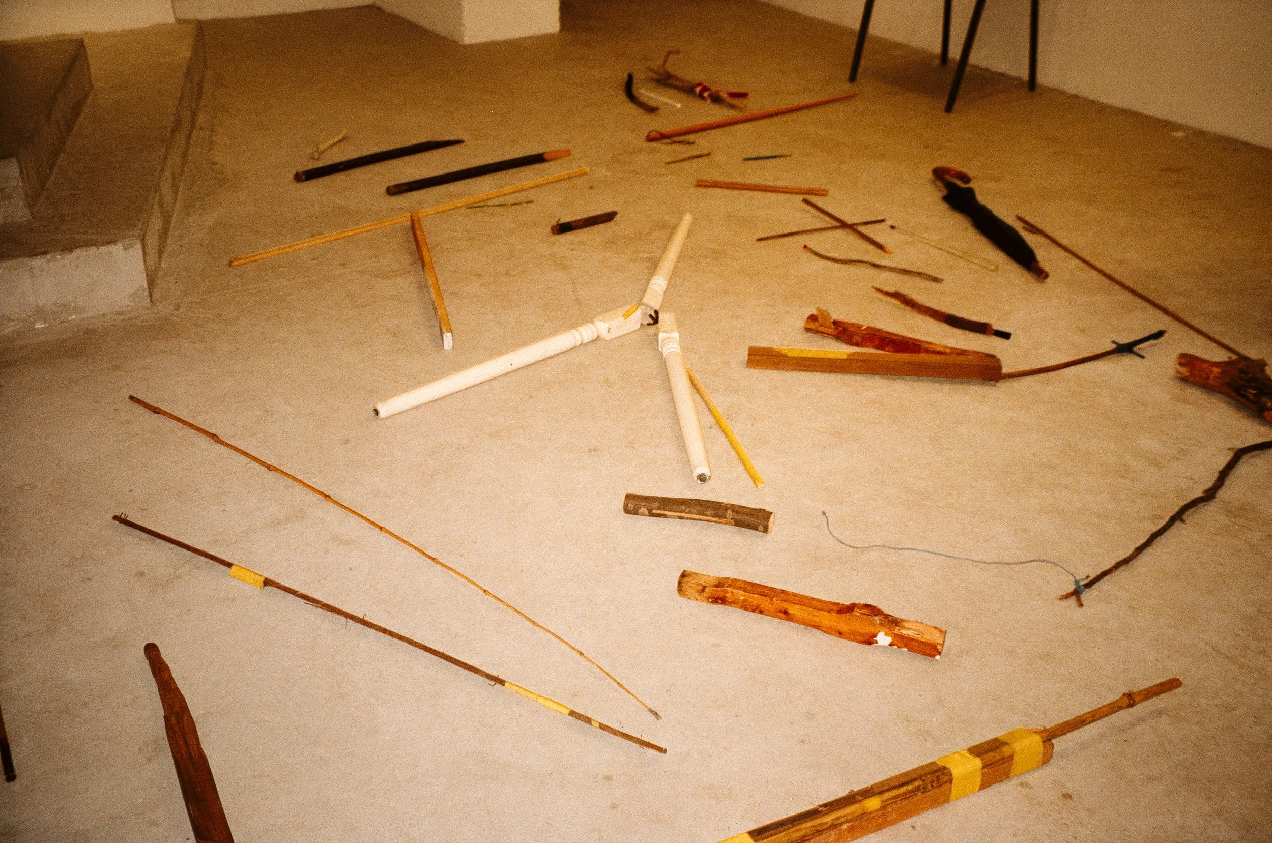 A room with various wooden sticks and pipes scattered on the floor, some painted yellow, white, or black, with a staircase on the left and a table with metal legs in the background.