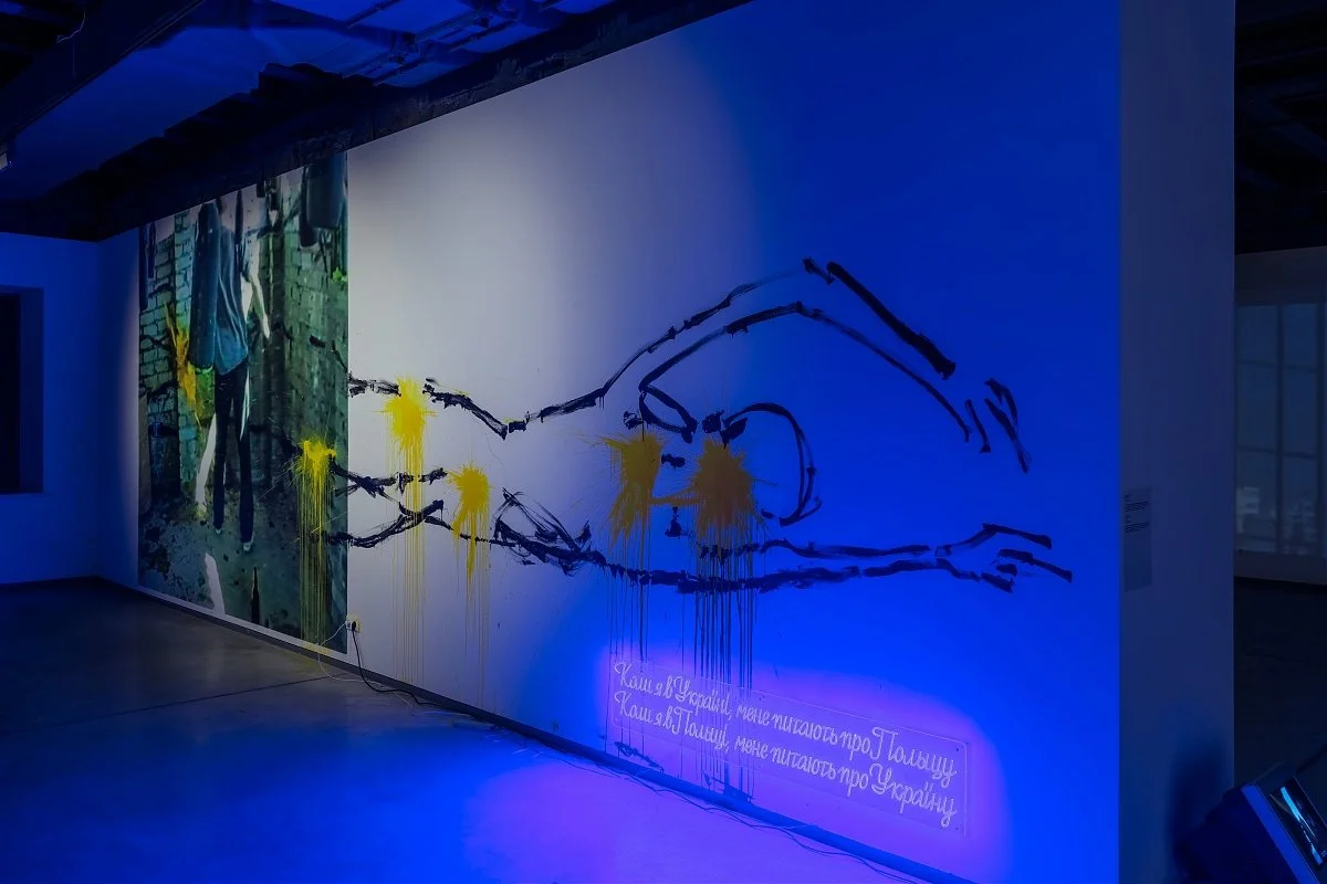 An art installation on a wall featuring a combination of a photograph, black painted outlines of a face, yellow paint splatters, and neon lights with Ukrainian text.
