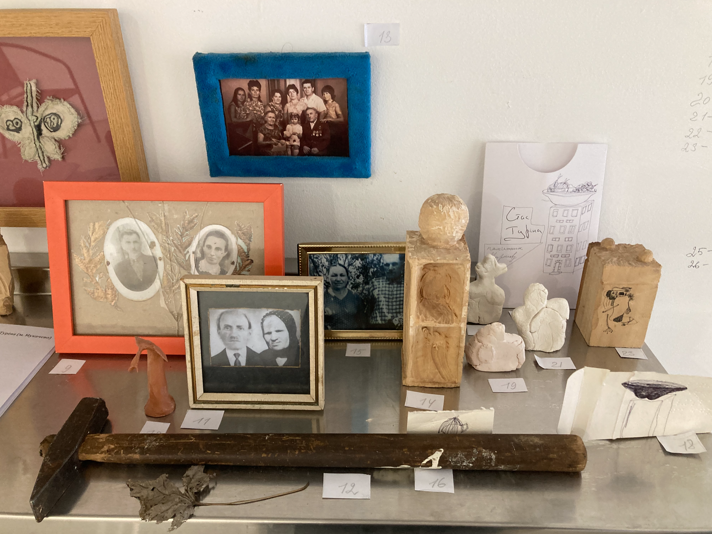 A display of various framed photographs, drawings, and small sculptures on a table and wall in a gallery or museum. The main photo shows a family portrait.