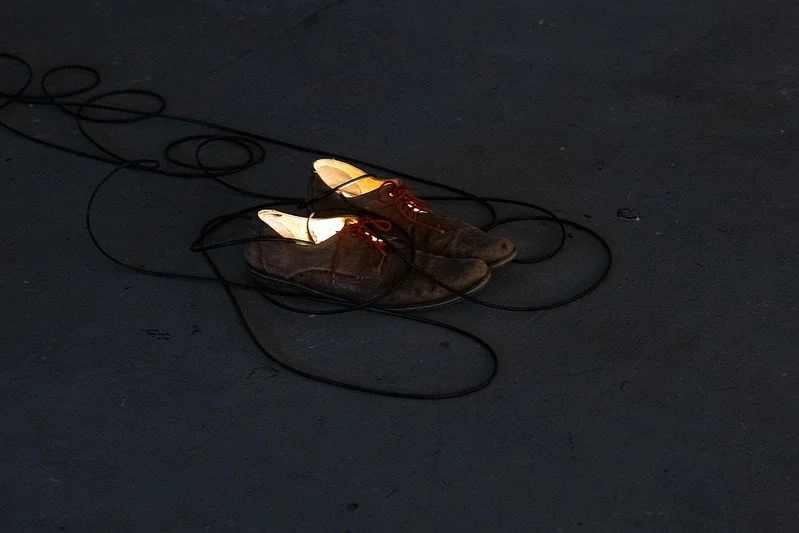 A pair of vintage brown shoes with white soles and red laces on a dark floor, surrounded by tangled black wires.