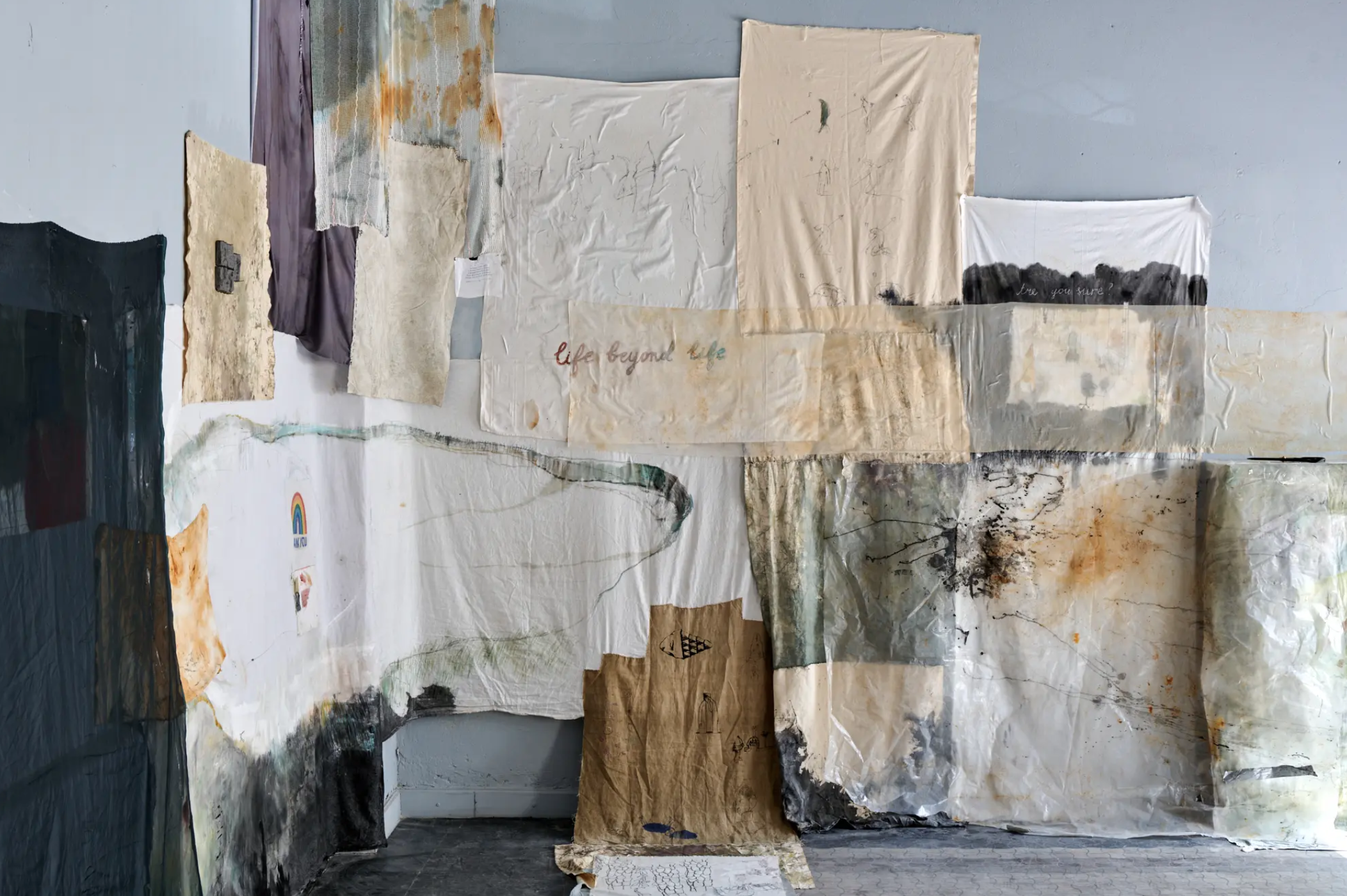 A gallery wall display of various textured and painted fabrics and papers, some with handwritten or printed text, and abstract or natural designs.