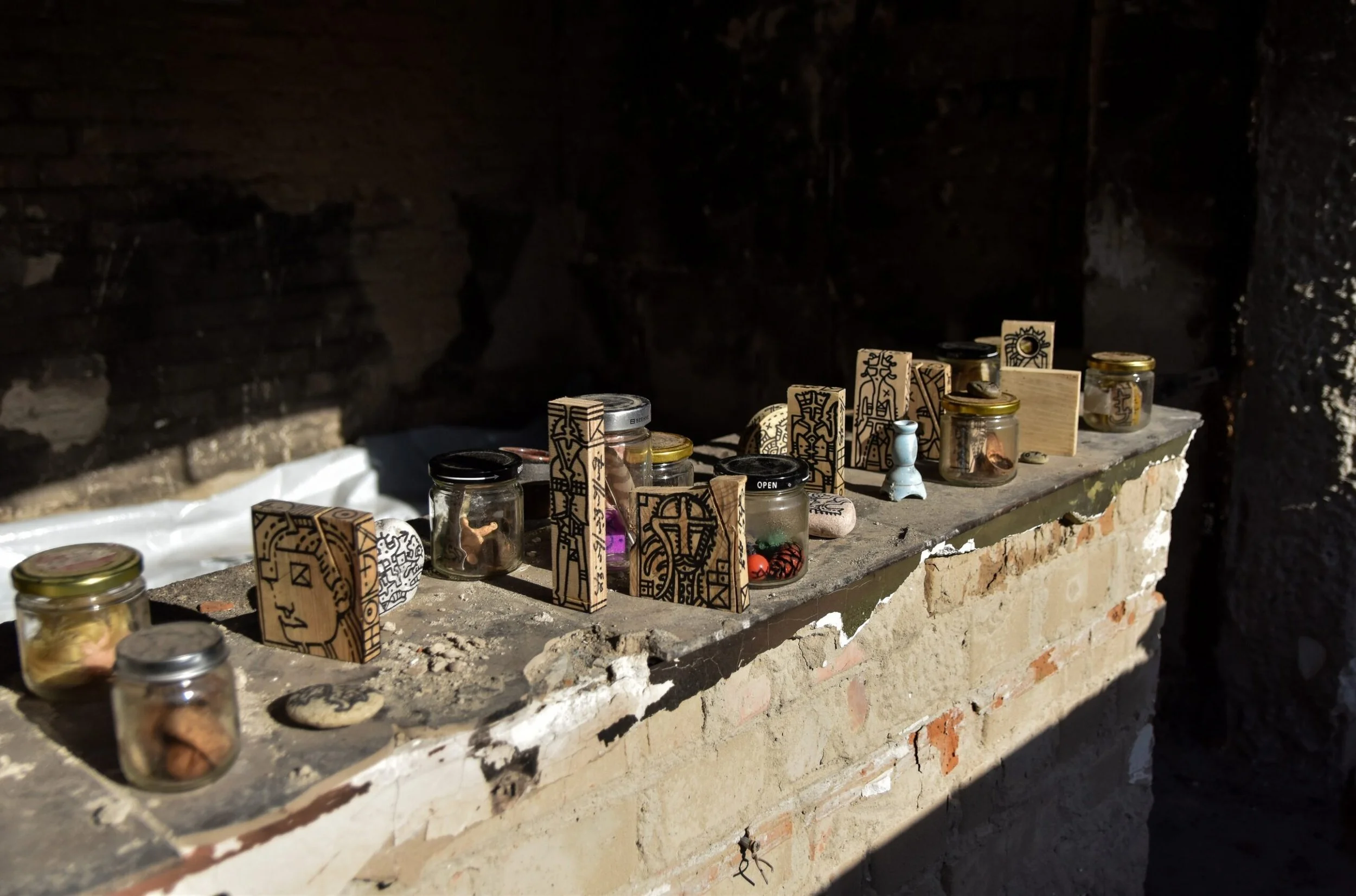 Small glass jars with various contents and decorative wooden stands on a stone ledge against a dark brick wall.