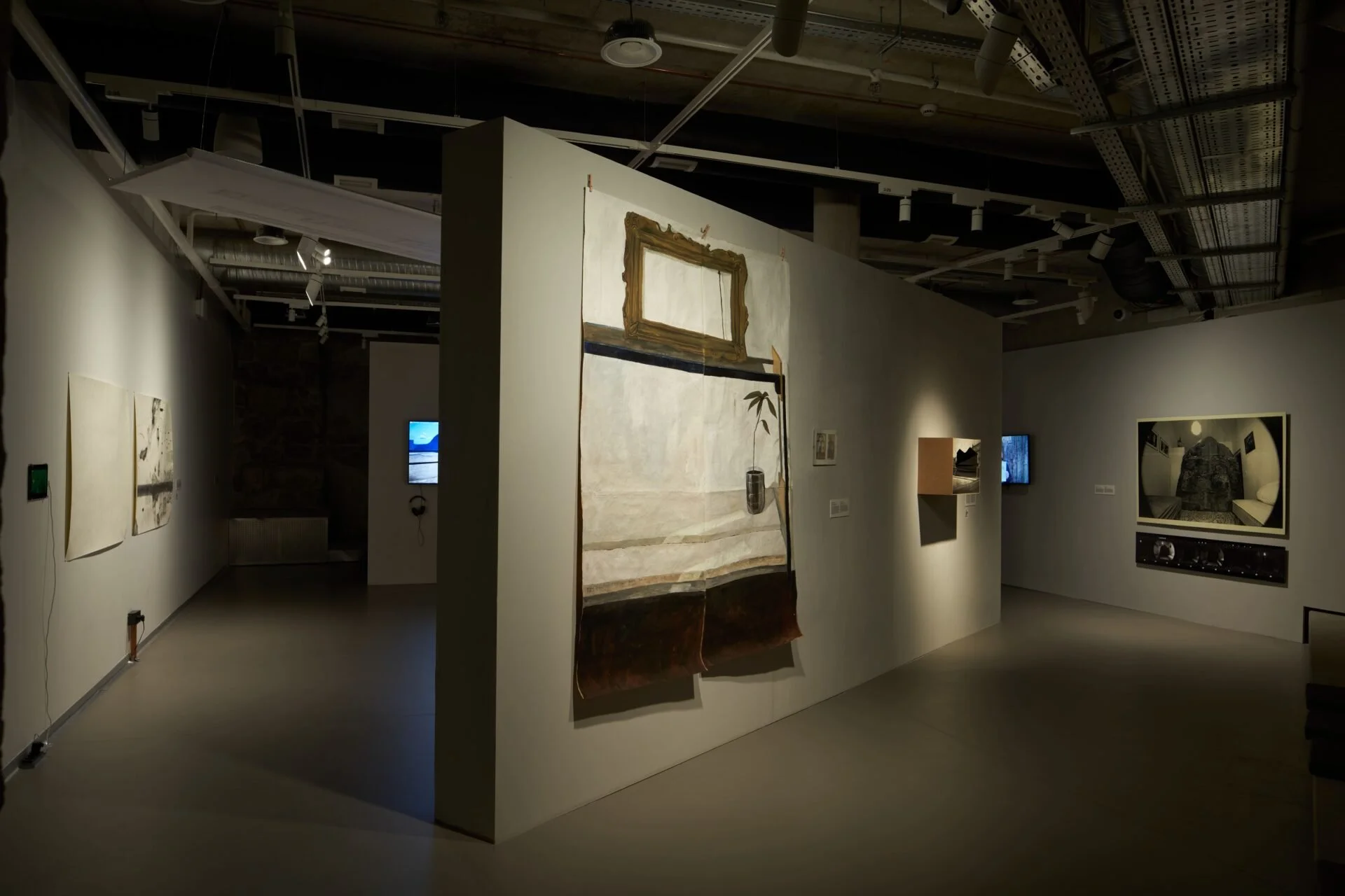 Art gallery with various paintings and digital screens on white walls, ceiling with exposed pipes, and track lighting.