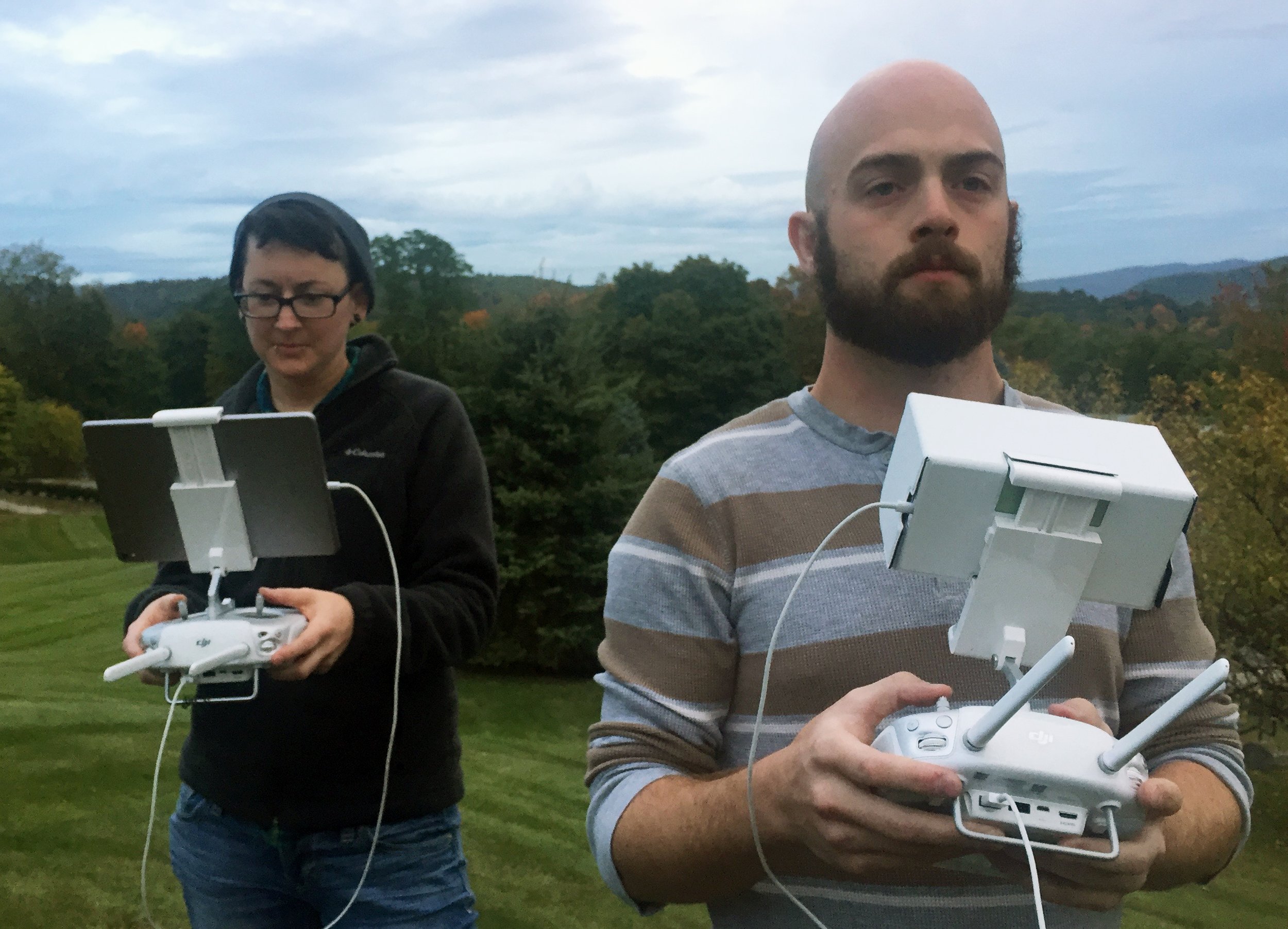 Two crew members standing outdoors on a grassy hillside, each holding a drone remote control with an attached monitor, flying a drone