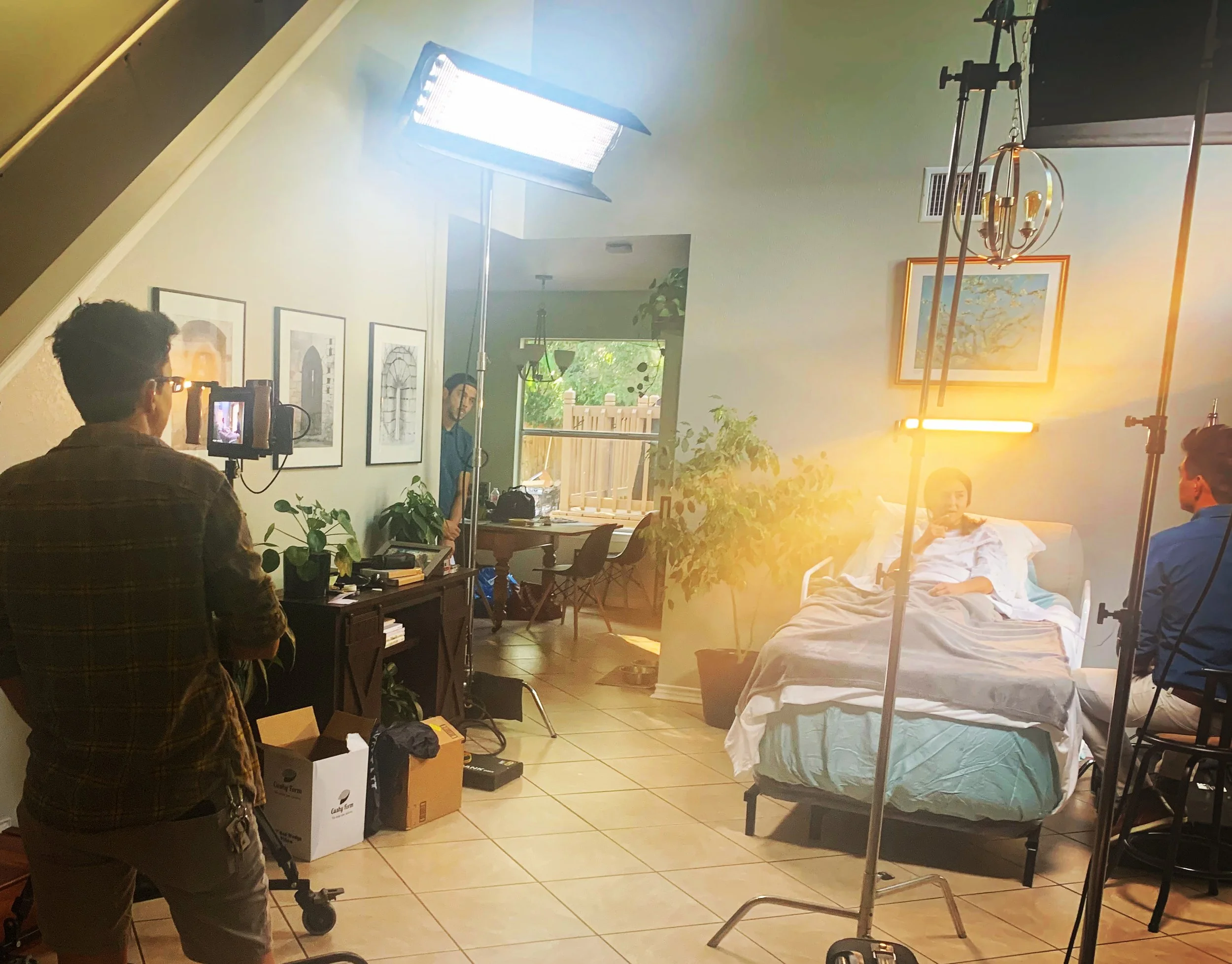 Behind-the-scenes photo of a video shoot, a crew member filming an actor lying in a hospital bed, with lighting equipment set up in a home interior