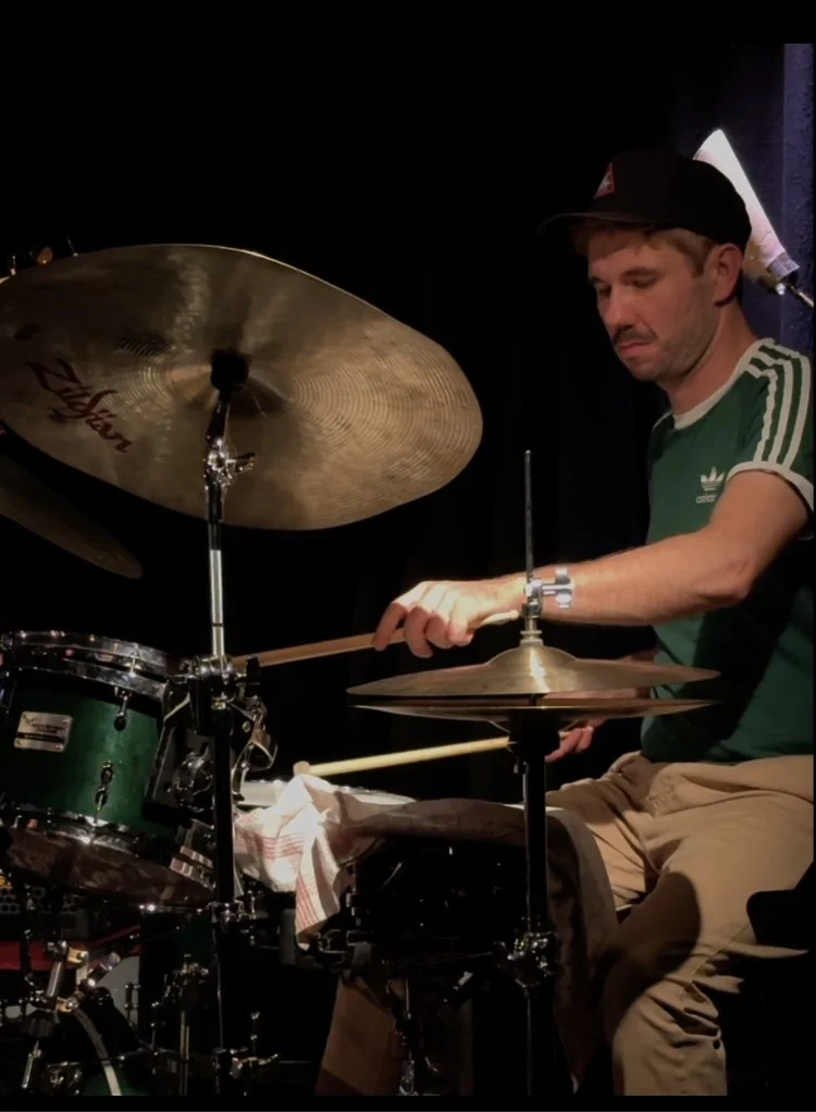 Max Autsch, Jazz Schlagzeug. Solo Musik. Ein Mann spielt Schlagzeug auf einer Bühne, trägt ein grünes T-Shirt, eine schwarze Mütze und sitzt vor einem schwarzen Hintergrund.