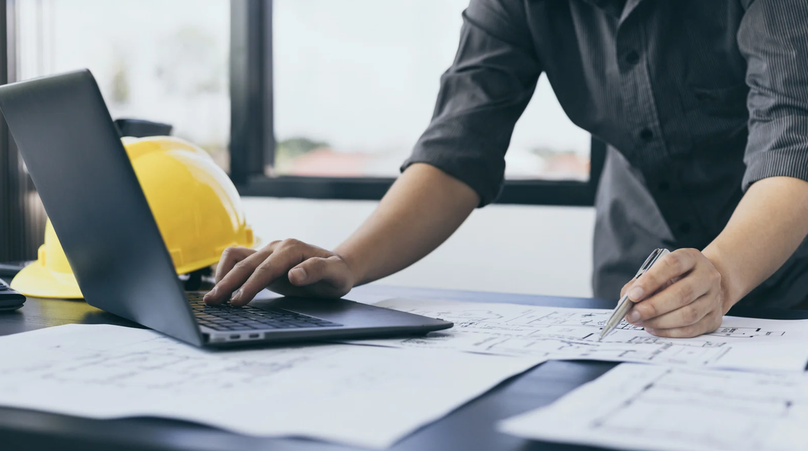 Person working at a desk with construction blueprints, a yellow safety helmet, and a laptop, in an office setting.