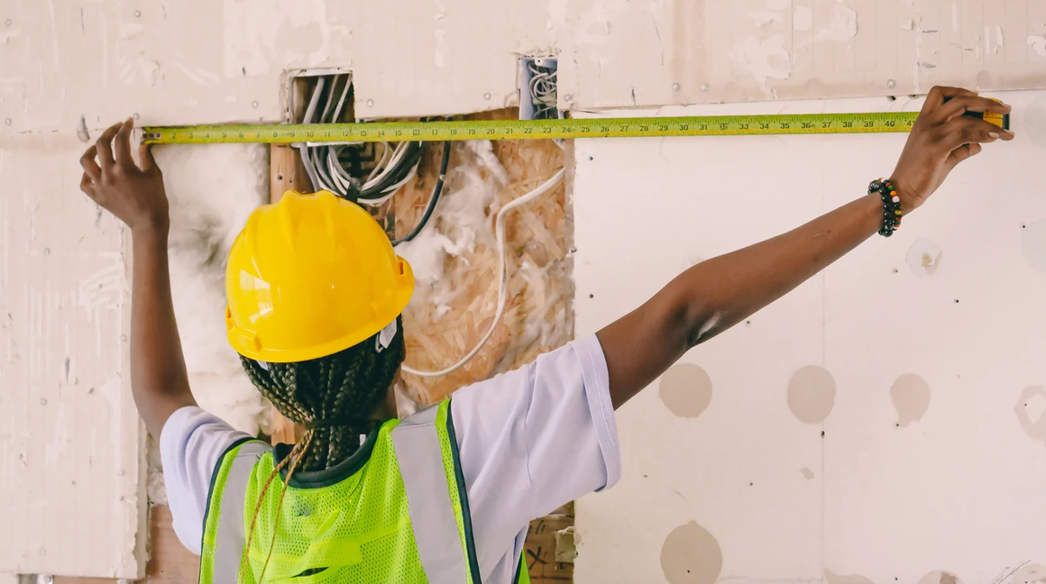 Worker in hard hat using a tape measure against a wall