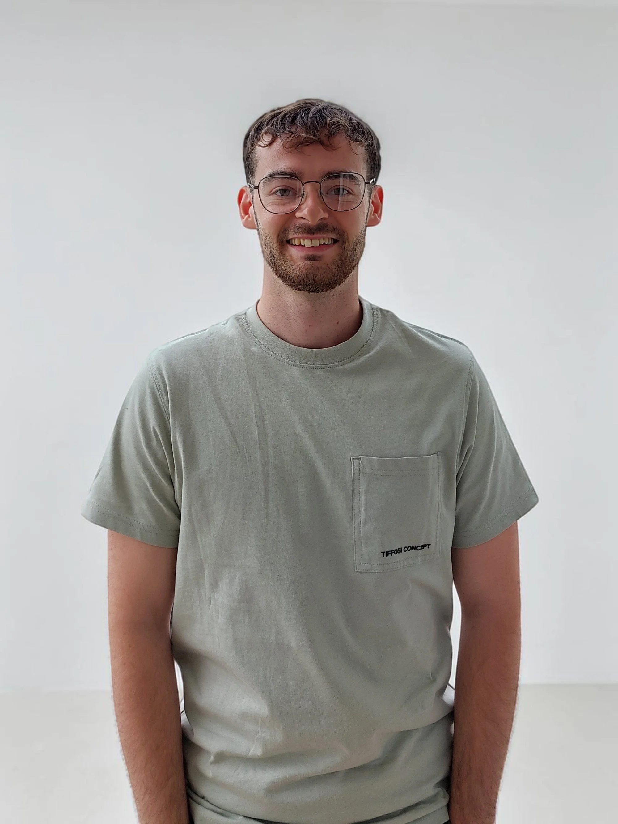 A smiling young man with glasses and a beard wearing a light gray T-shirt, standing against a plain white background.