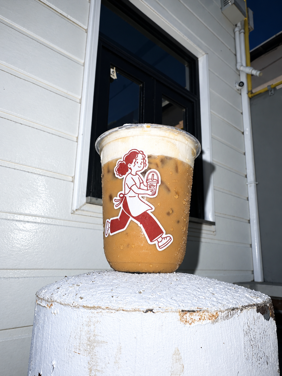 Vaso de latte en las rocas con coldfoam  de vainilla con unaa ilustración de la monita de Casa Cosmopolita que corre y sostiene un vaso con bebida y popote, en un fondo de pared blanca y ventana.