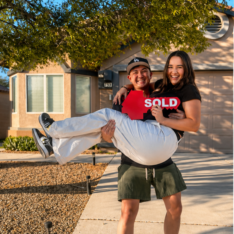A young man holding a young woman who is holding a red 'SOLD' sign in front of a house, celebrating a home sale.
