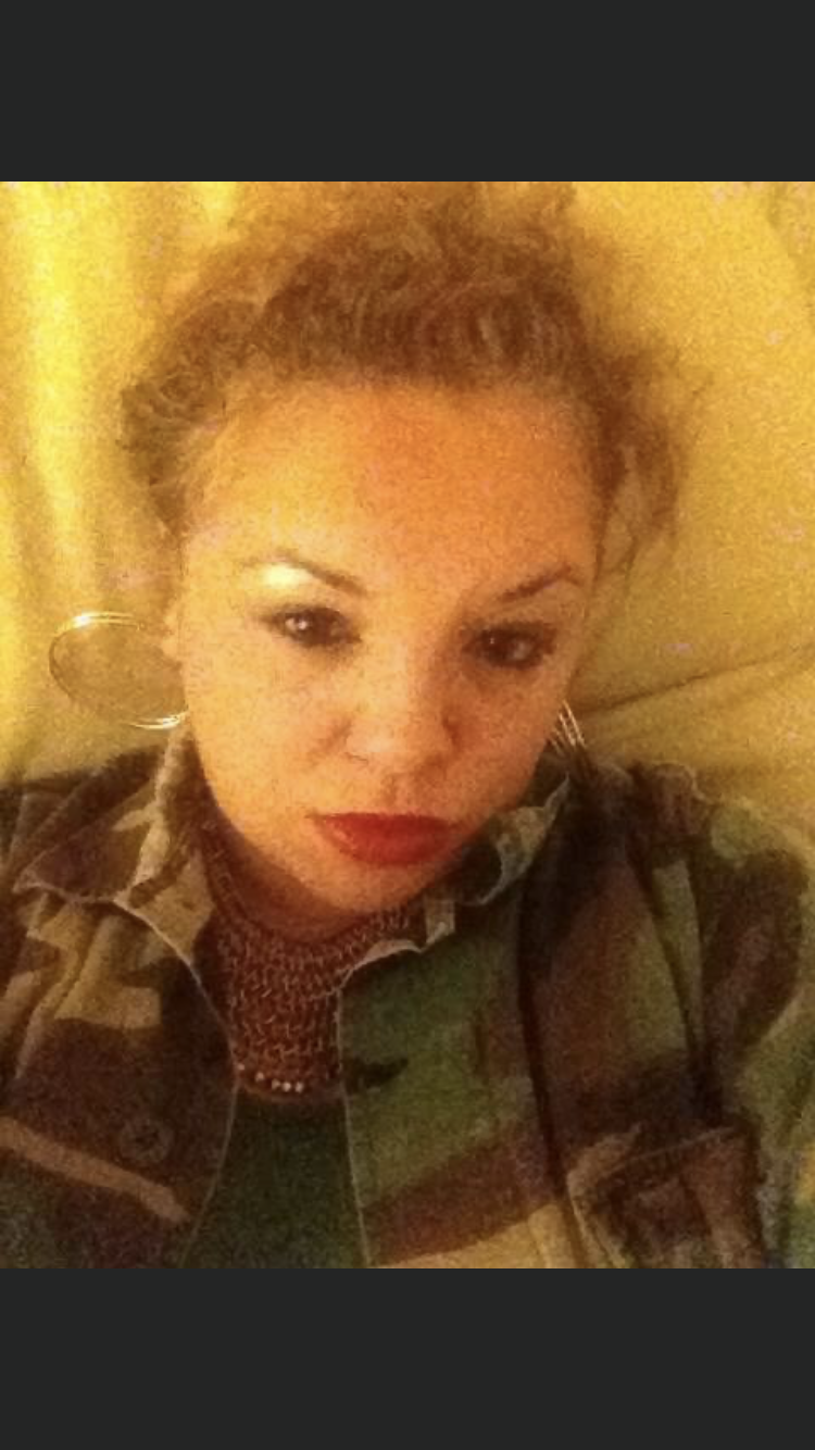 A woman with curly hair and gold hoop earrings is taking a selfie. She is wearing a camouflage military jacket and a patterned top, with makeup including lipstick and eye makeup. She is lying down against a pillow or headboard with a yellowish lighting.