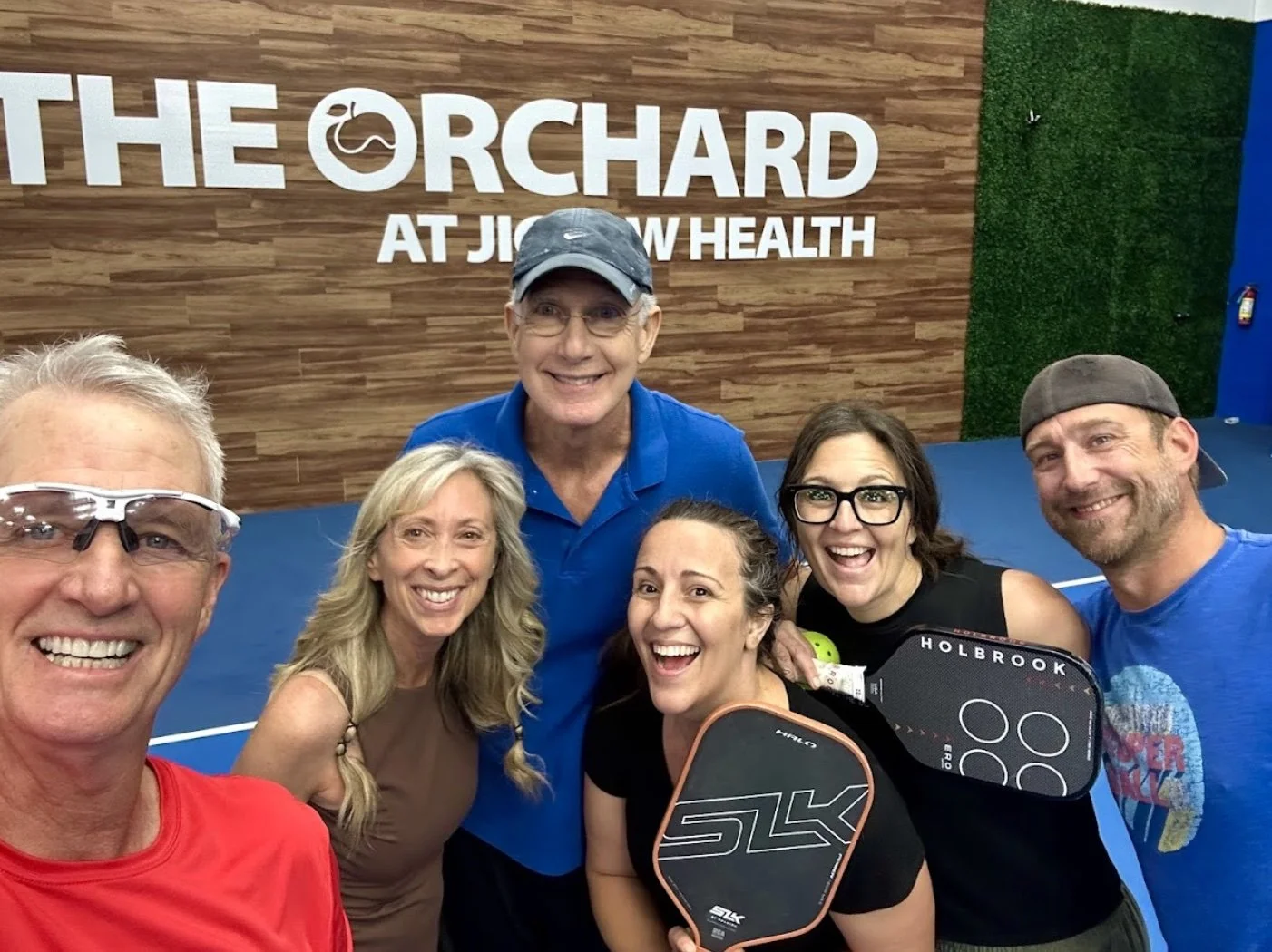 Group of six people smiling for a selfie on an indoor paddle tennis court, holding paddles, with a wooden and green wall background that has the logo and text 'The Orchard at Jowsey Health'.