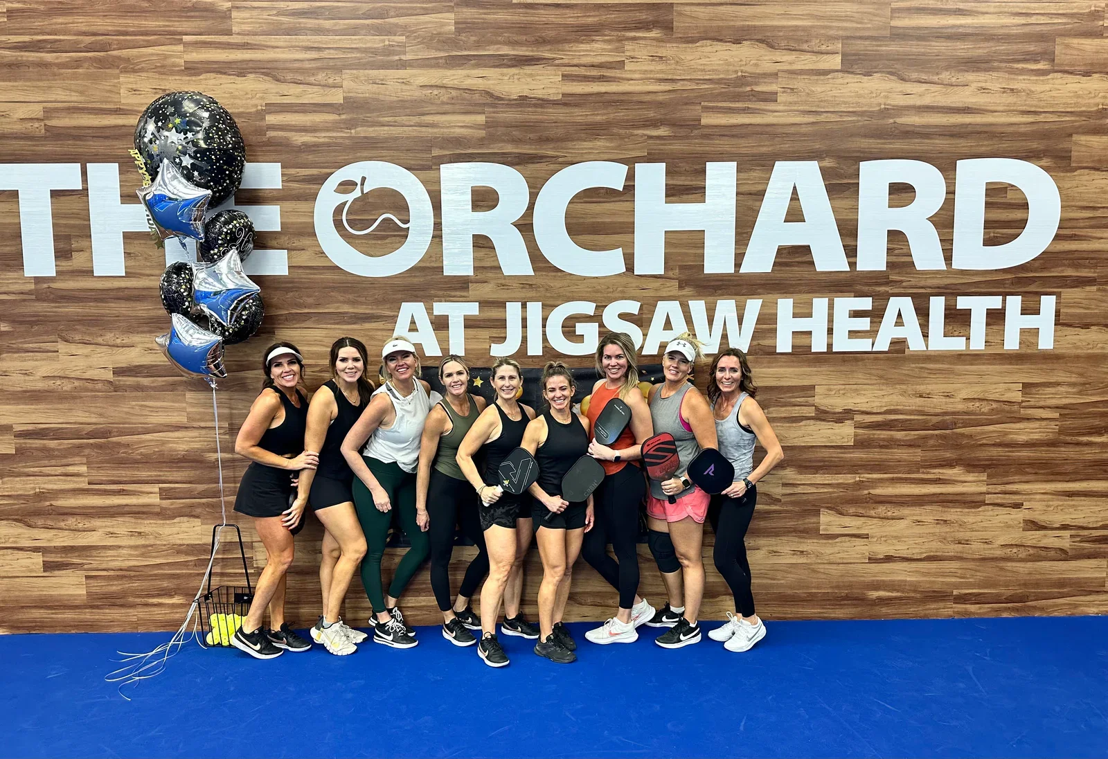 A group of women in workout attire standing in front of a wall with large sign that reads 'THE ORCHARD AT JIGSAW HEALTH', holding paddleboarding paddles, with balloons nearby.