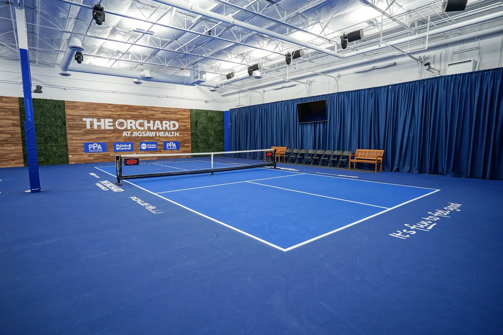Indoor pickleball court with blue flooring and a net, surrounded by blue curtains and seating, with signage that says 'The Orchard at Jigsaw Health' and logos for pickleball associations.