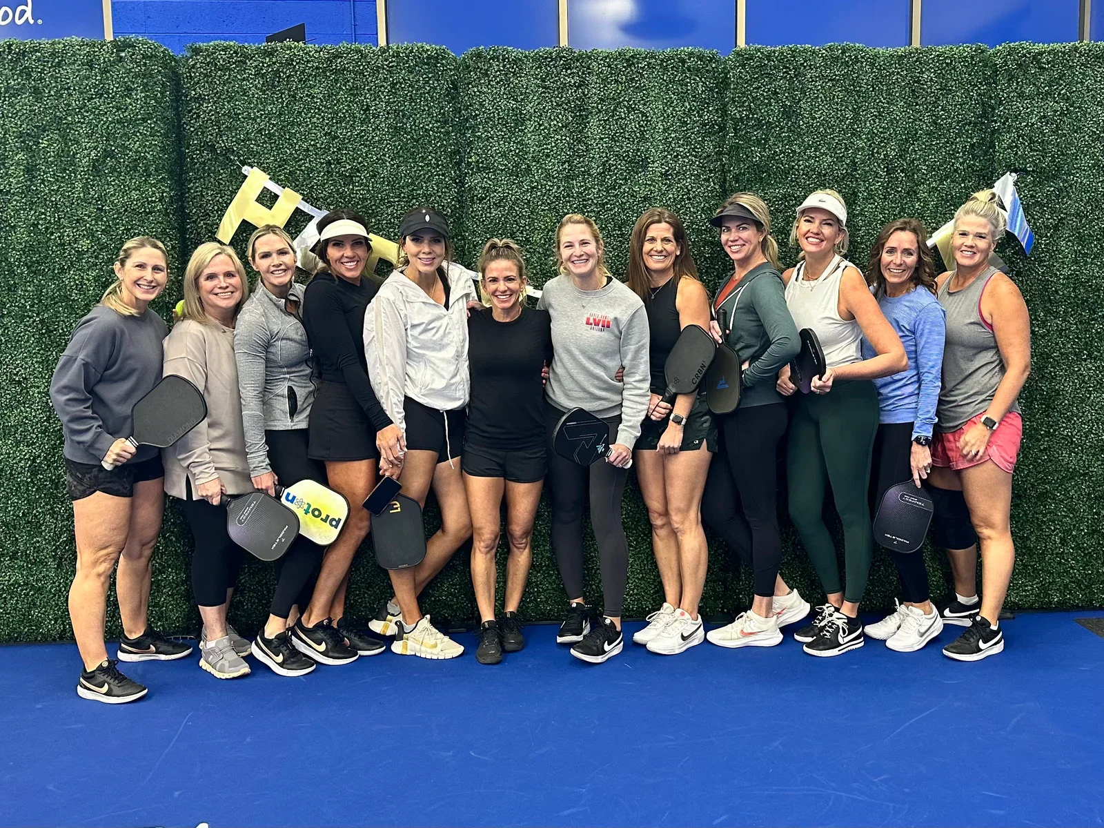 Group of women in athletic clothing holding pickleball paddles, standing in front of a green hedge wall at an indoor sports facility.