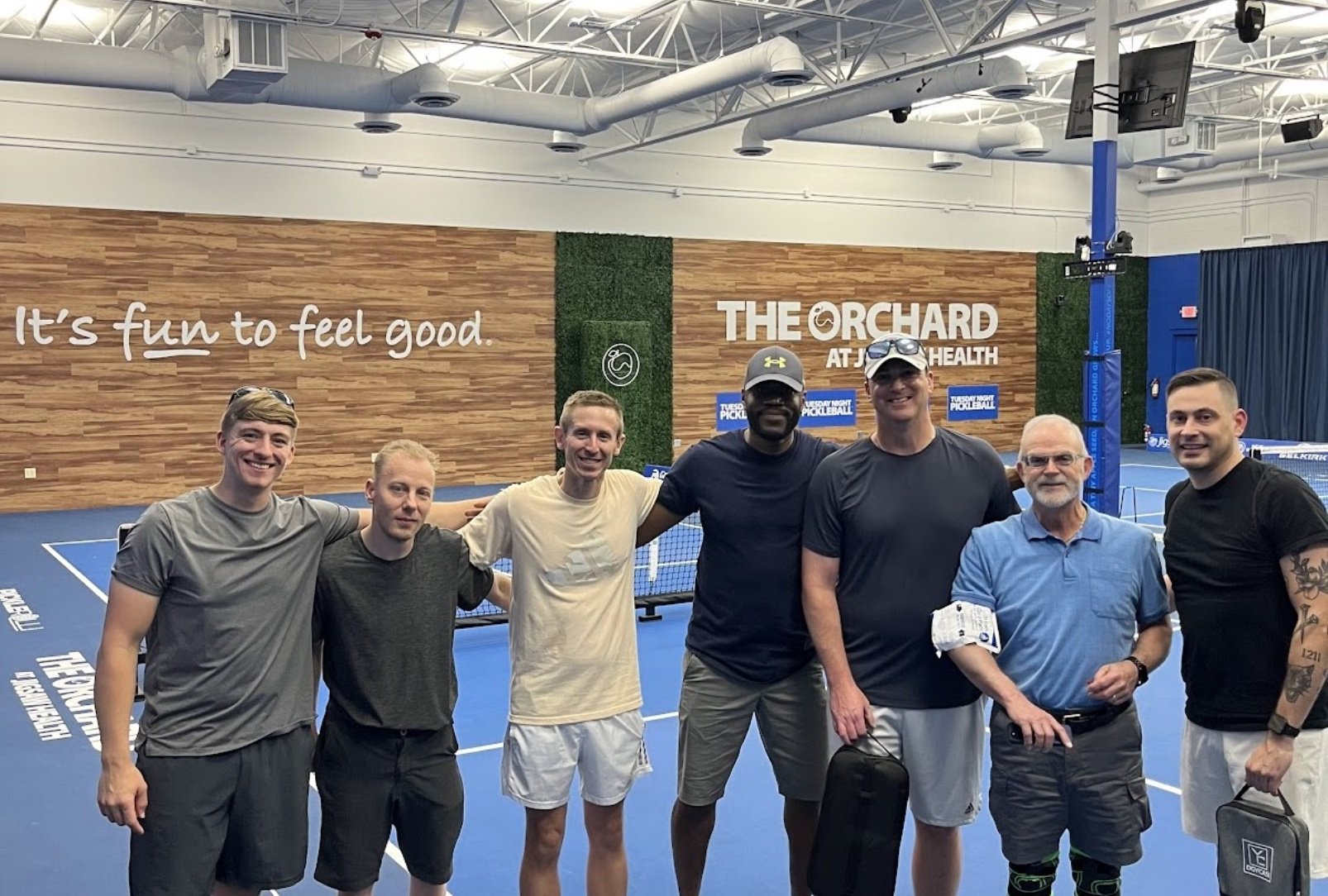 A group of seven men standing together on a blue indoor pickleball court, smiling, with a wall behind them that has the words "It's fun to feel good" and "The Orchard at JCC Health." The group appears to be at a pickleball event, with some dressed in