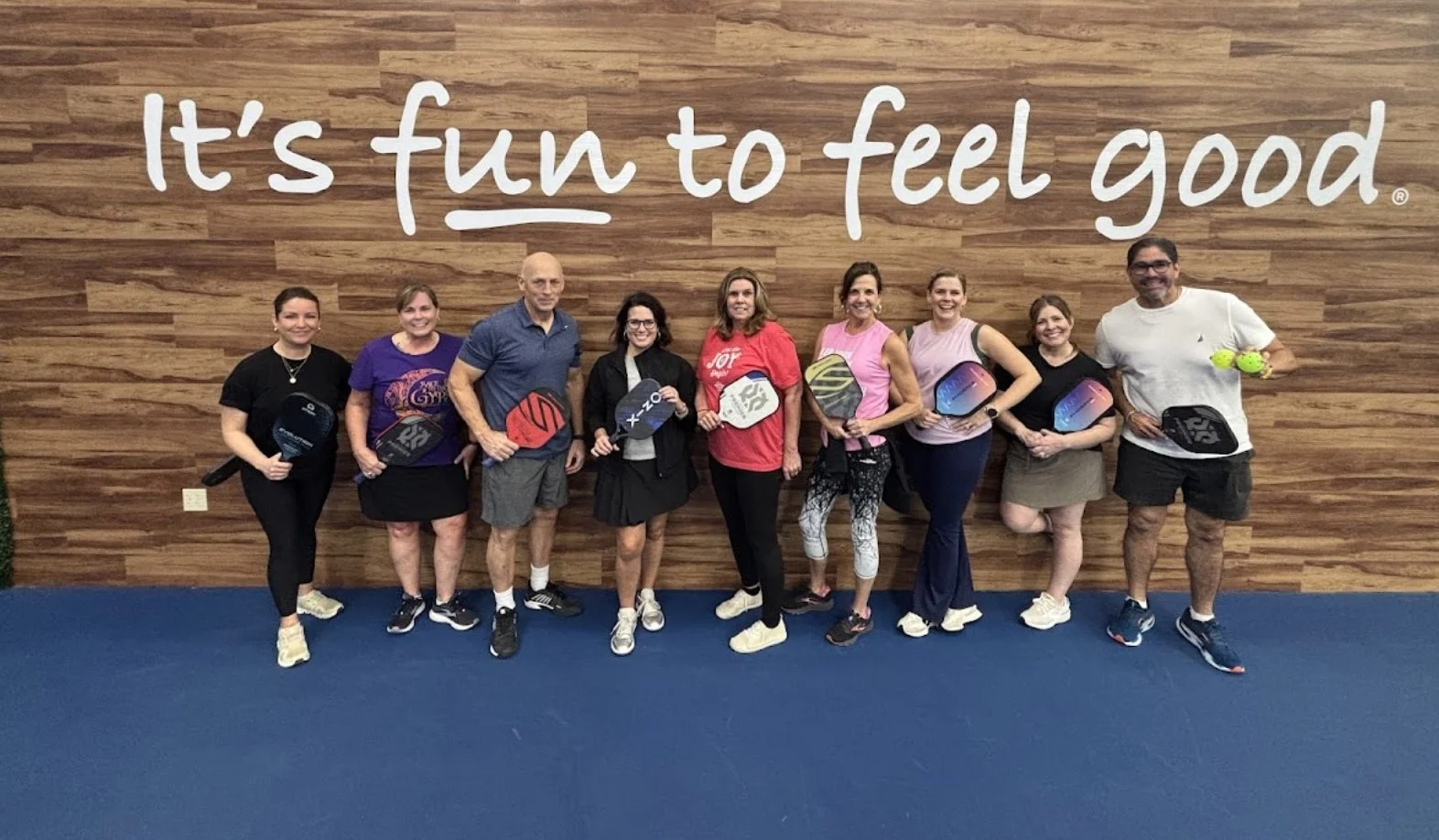 Group of nine people standing indoors in front of a wooden wall with the phrase "It's fun to feel good" written on it, holding pickleball paddles and wearing casual sportswear.