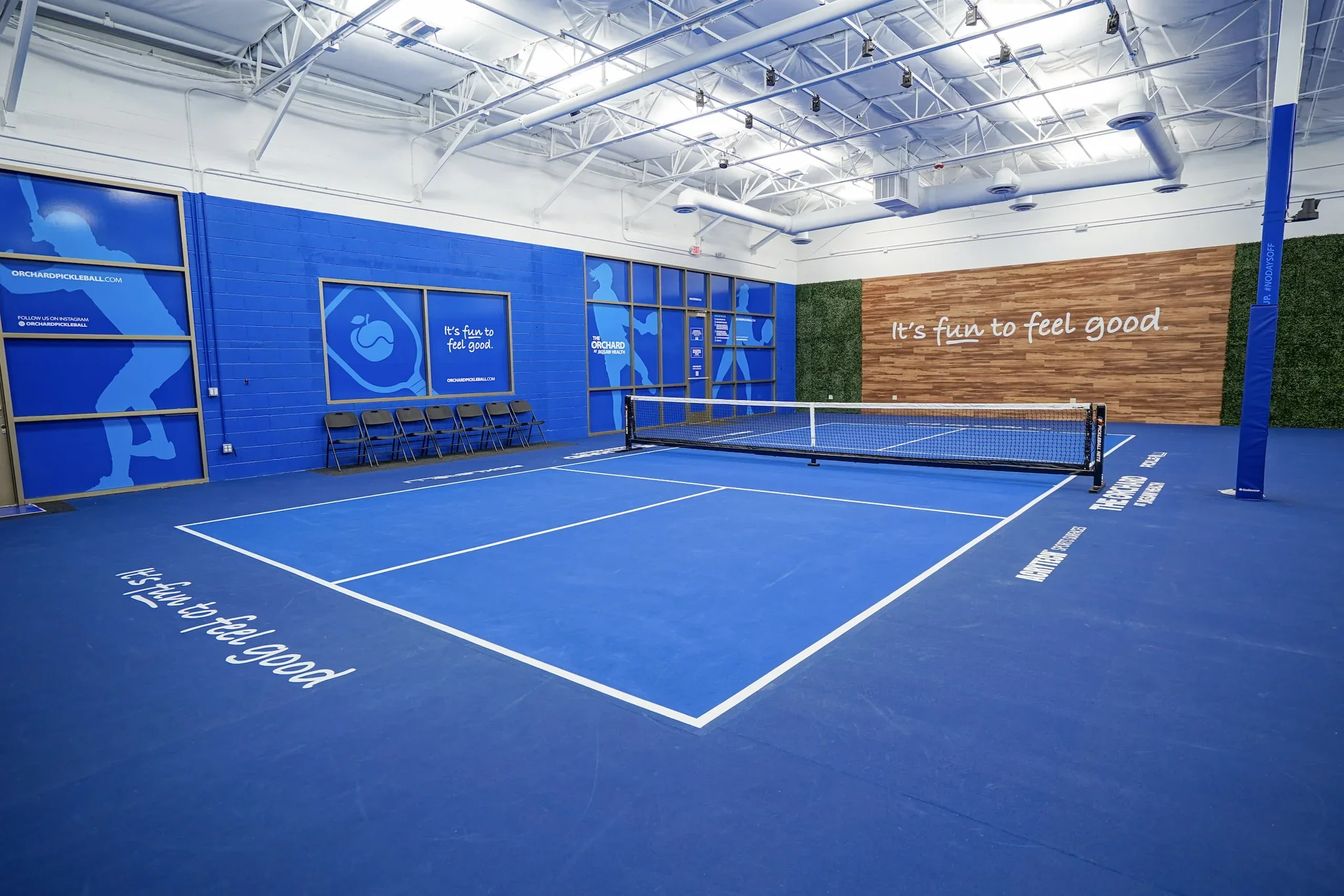 Indoor pickleball court with blue flooring, white markings, and a net in the middle. The walls have blue and green accents, with motivational messages and graphics related to pickleball.