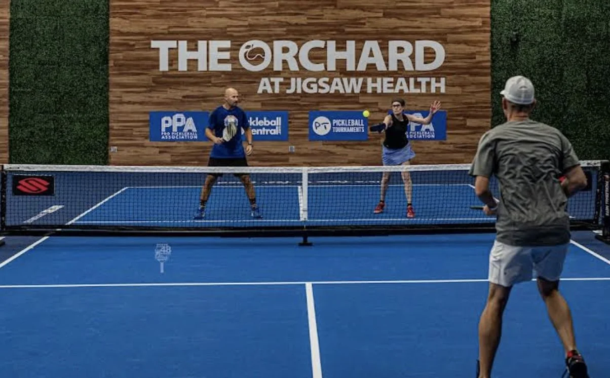 People playing pickleball on an outdoor court at The Orchard at Jigsaw Health, with a wooden wall and green hedges in the background.