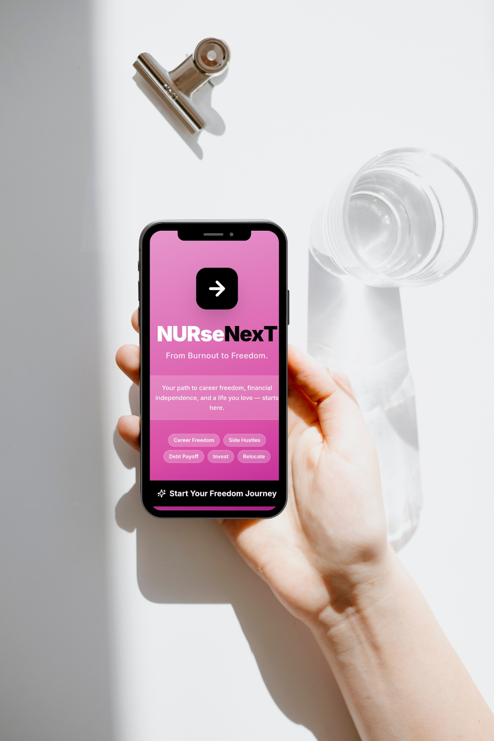 A person holding a smartphone displaying the NURseNextT program app on a pink background, with a glass of water, a metallic clip, and a white cloth on a white surface.