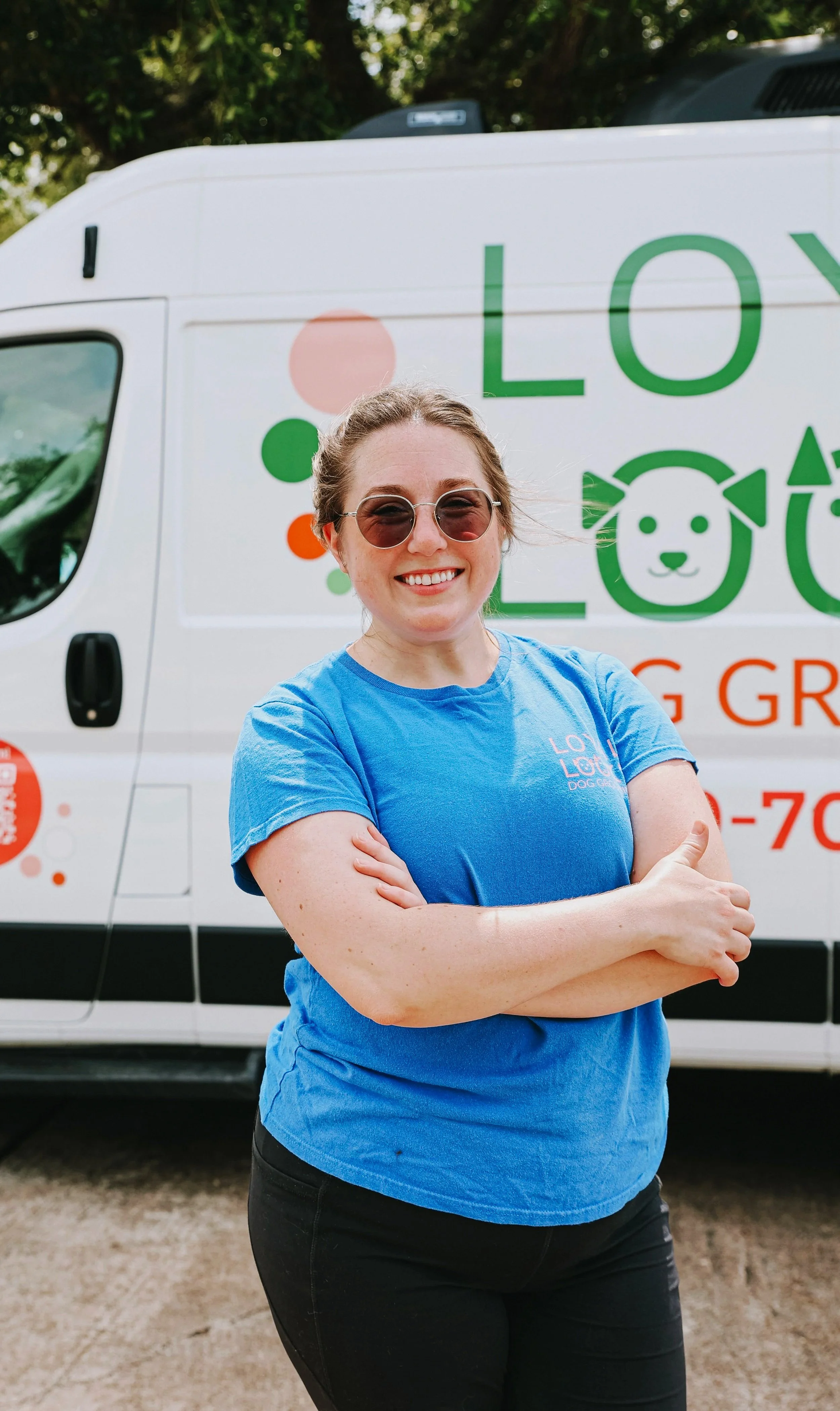 A smiling woman wearing sunglasses and a blue T-shirt standing with her arms crossed in front of a white animal rescue van with green and orange lettering and graphics.