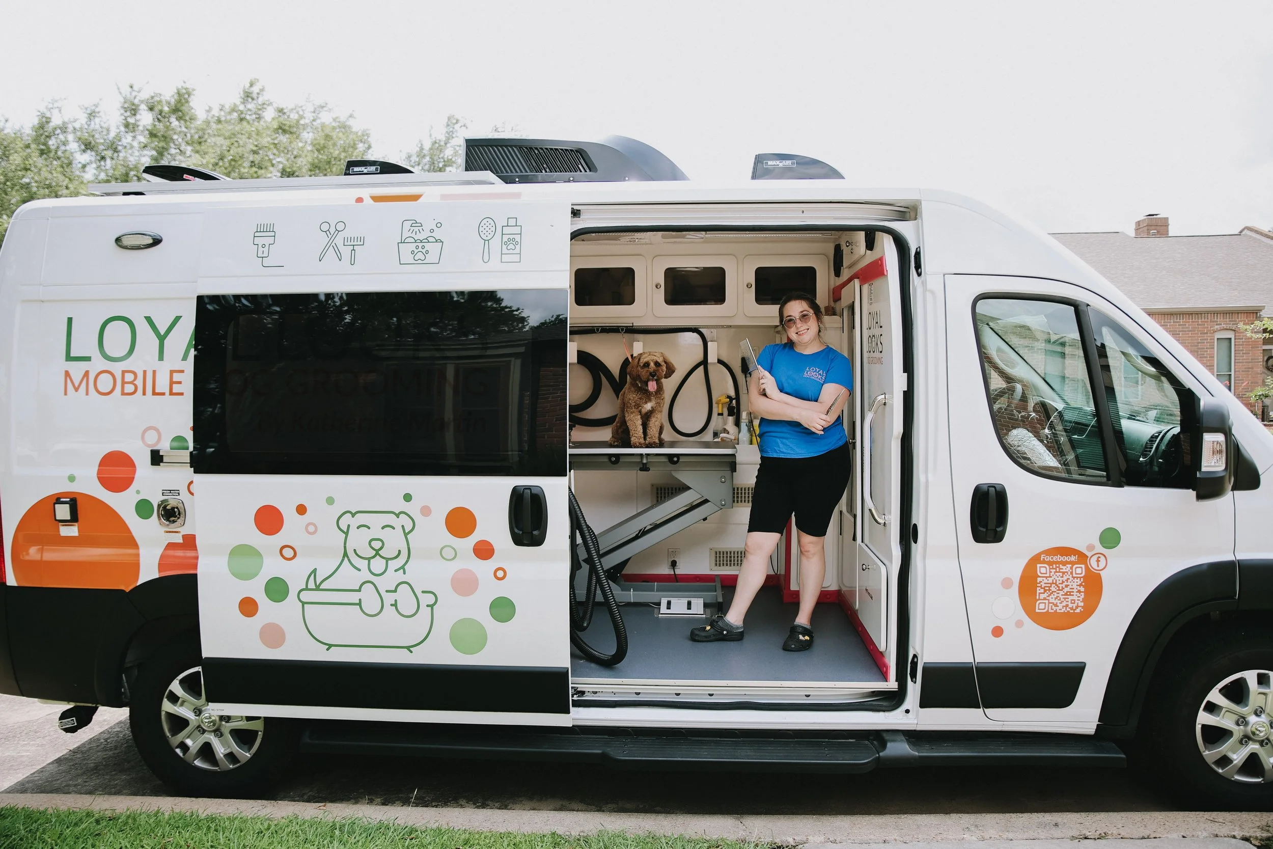 A woman in a blue T-shirt and black shorts standing inside a mobile pet grooming van with a small brown dog on the grooming table. The van is decorated with colorful dog-related graphics and icons, and the background shows a residential neighborhood with trees and houses.