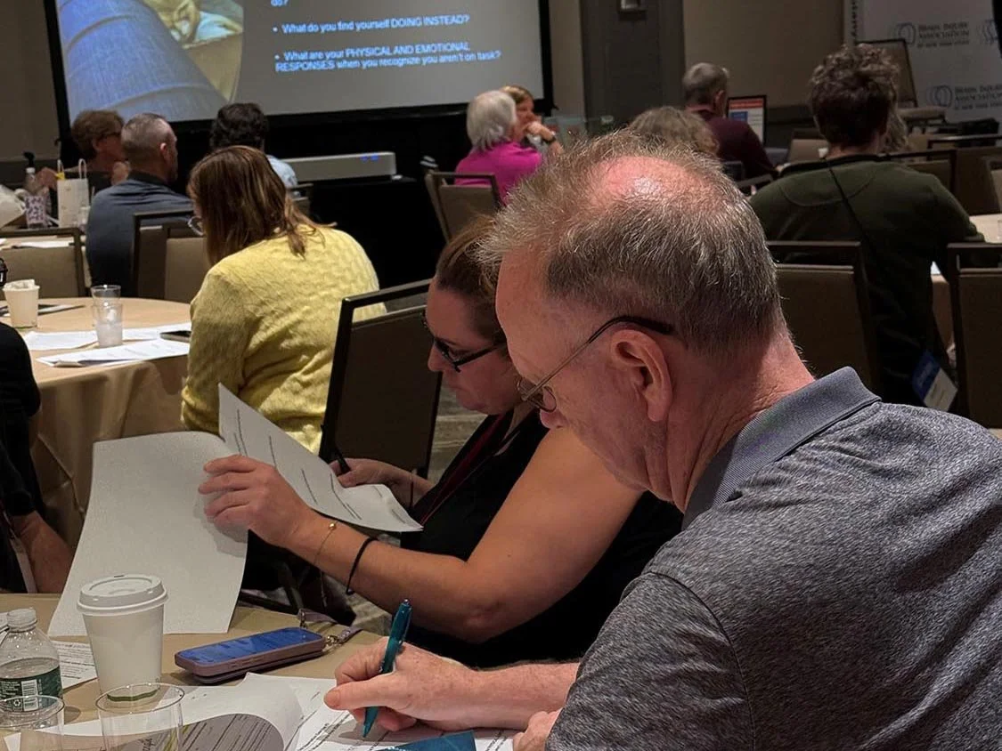 Individuals sitting at a conference looking at a projector screen taking notes