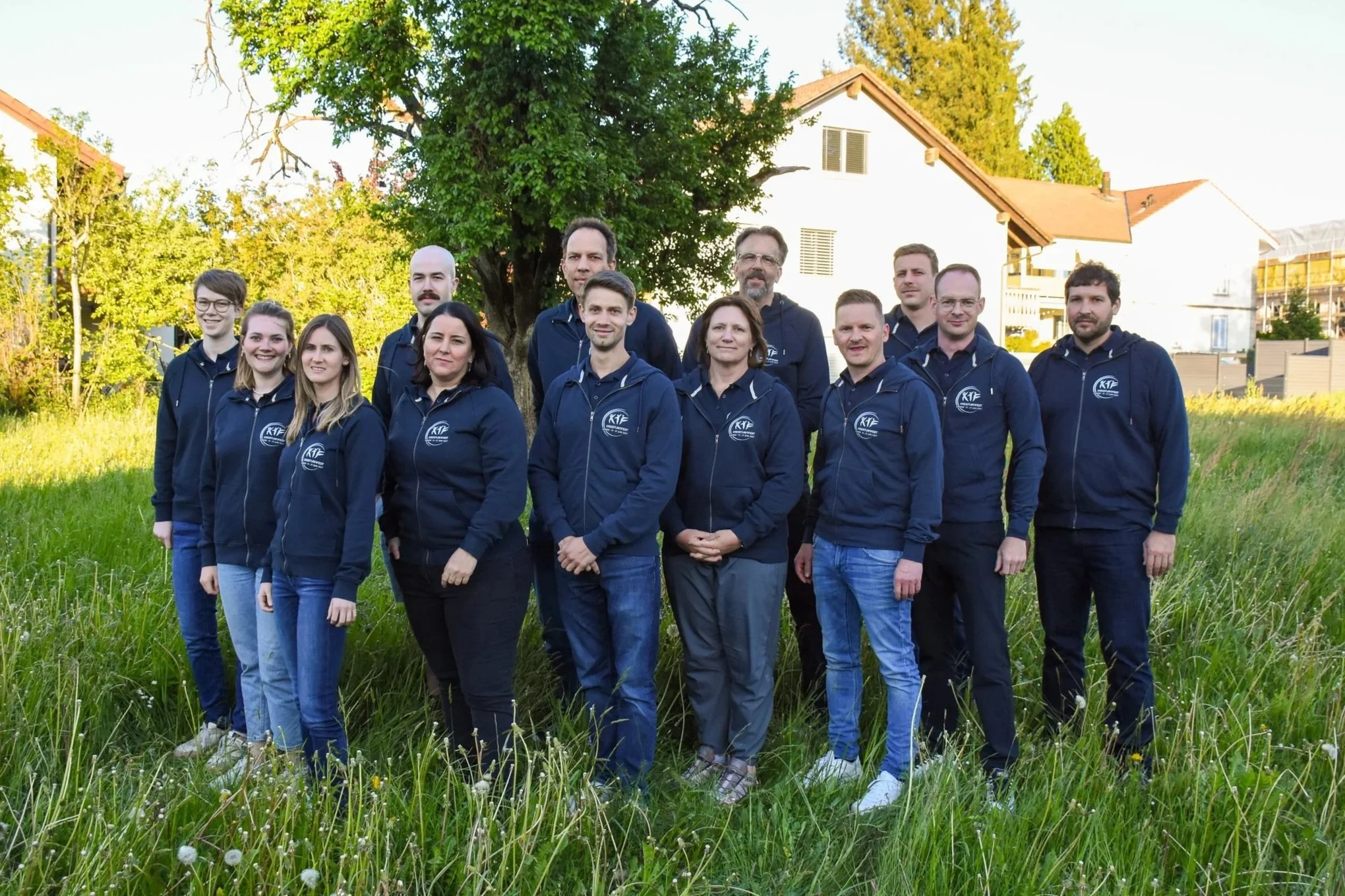 Eine Gruppe von 13 Menschen in dunklen Jacken mit einem Logo steht im Freien vor einem Baum und einem Haus. Es ist Tag und das Wetter ist sonnig.
