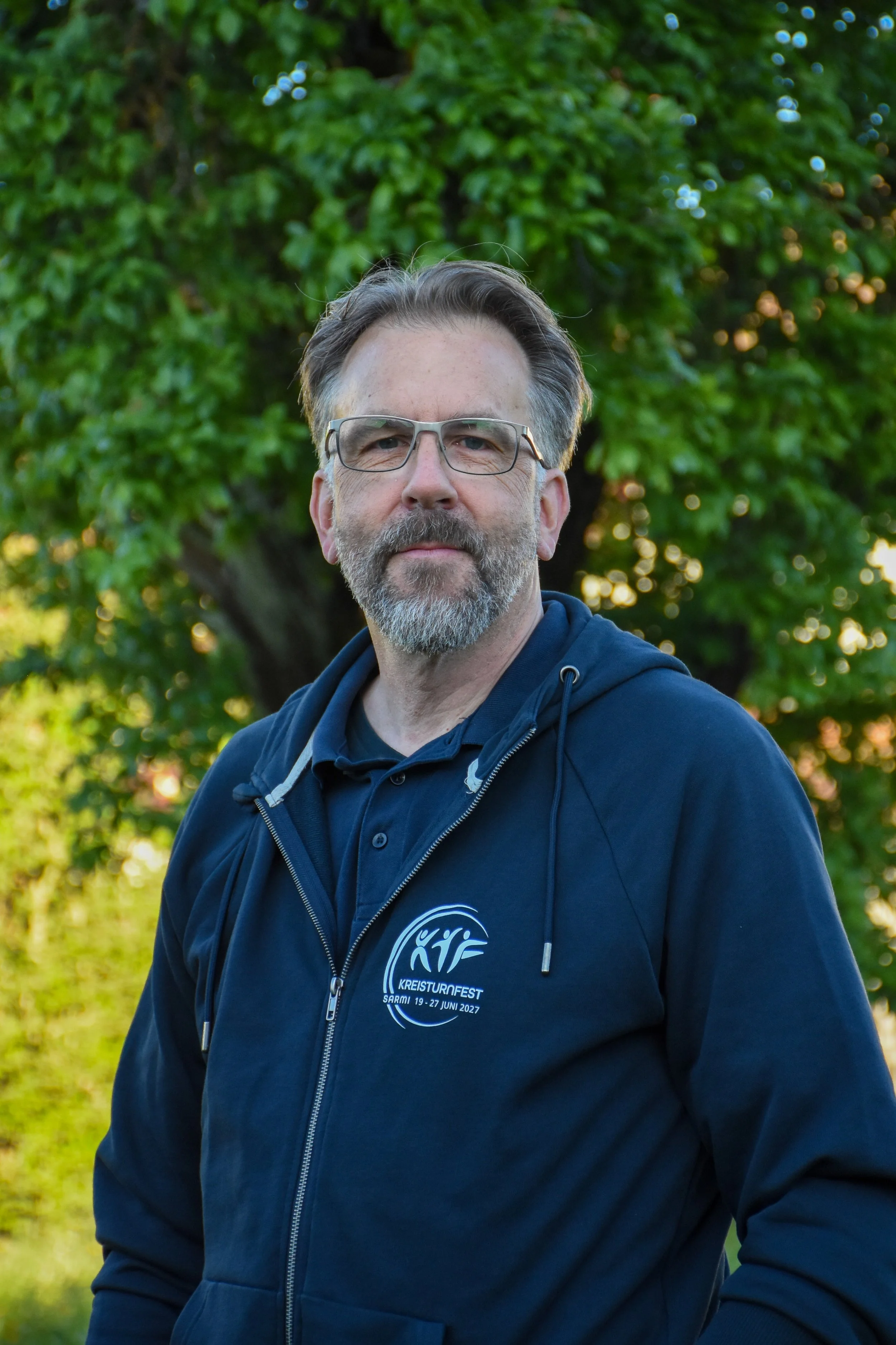 Ein Mann mit Brille, grauem Bart und dunklem Haarschopf steht im Freien vor einem Baum mit grünen Blättern. Er trägt einen blauen Hoodie mit einem Logo und Text, der auf eine Veranstaltung im Juni 2027 hinweist.