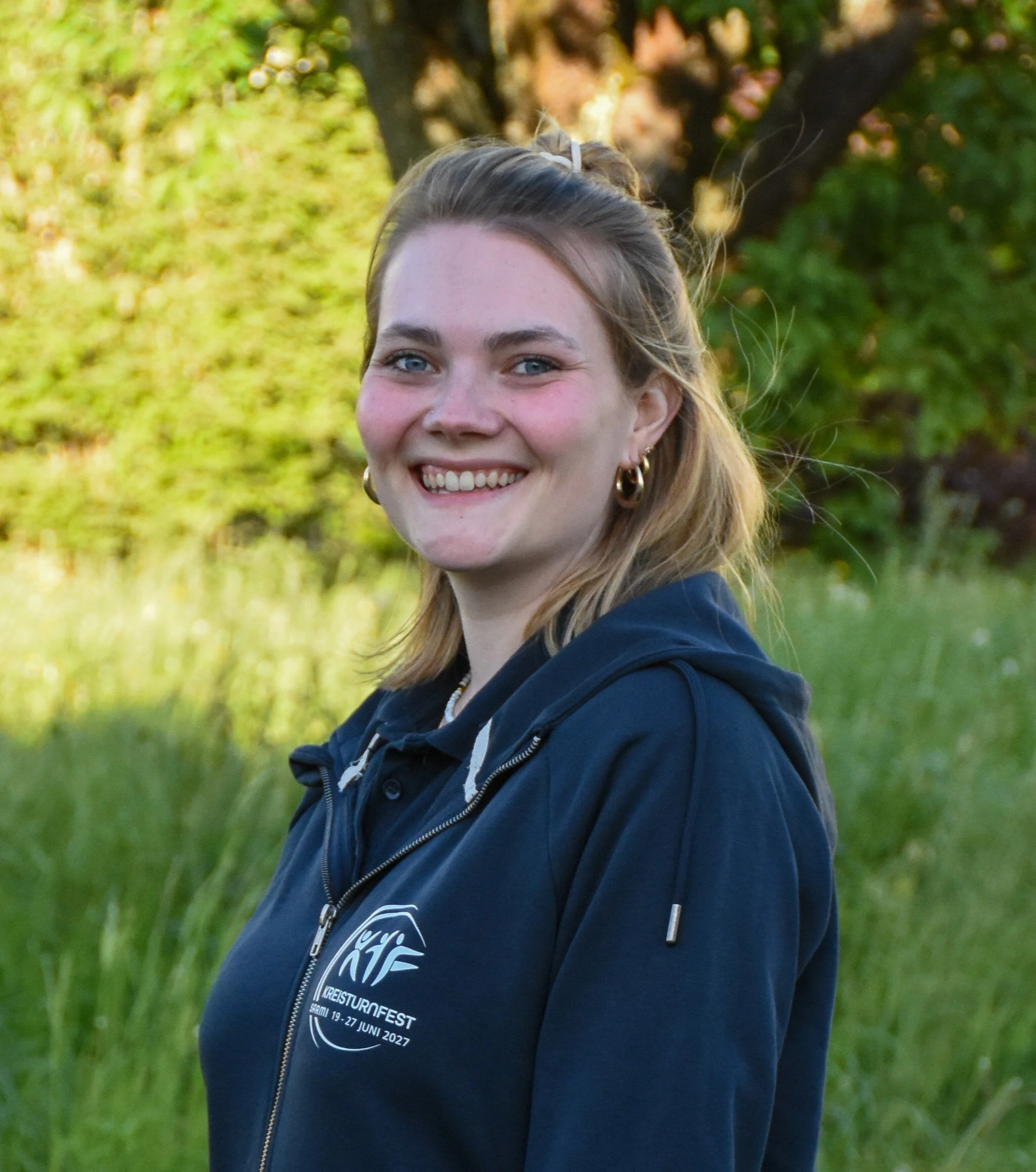 Junge Frau mit blonden Haaren, blauen Augen, goldenen Ohrringen und einem blauen Hoodie, lächelnd im Freien vor grünen Bäumen bei Sonnenlicht.