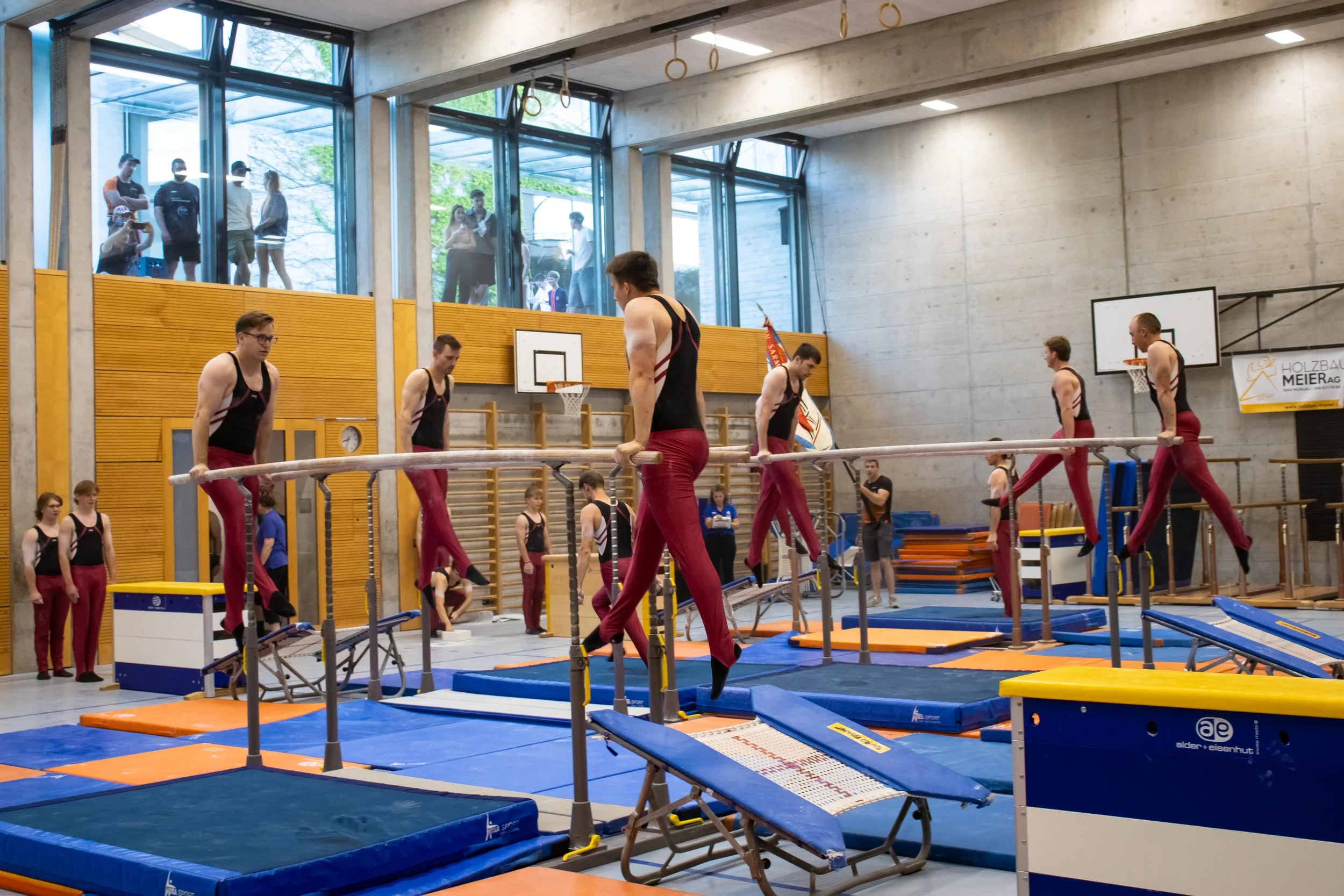 Sporthalle mit Turngeräten und mehreren jungen männlichen Turnern, einige auf der Schwebebank, andere im Hintergrund beim Training, mit Zuschauern außerhalb der Glasfenster.