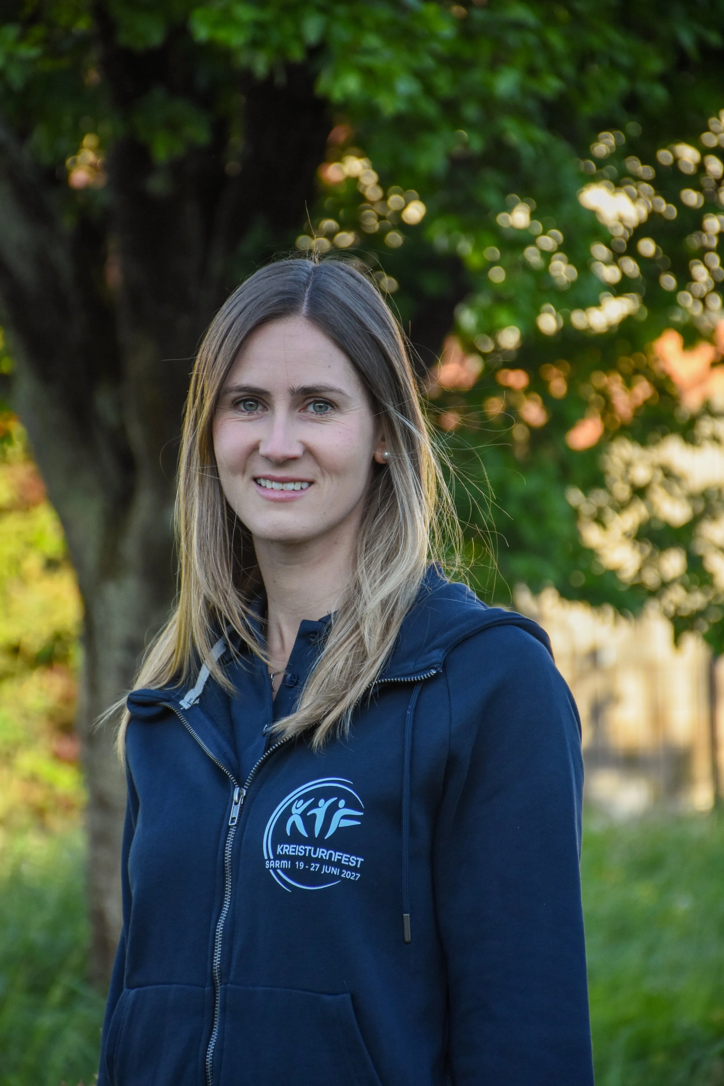 Junge Frau mit blonden Haaren im Freien, trägt eine dunkelblaue Jacke mit Logo und Text für das KREISTURNFEST vom Juni 2027, im Hintergrund Bäume und bunte Herbstblätter.