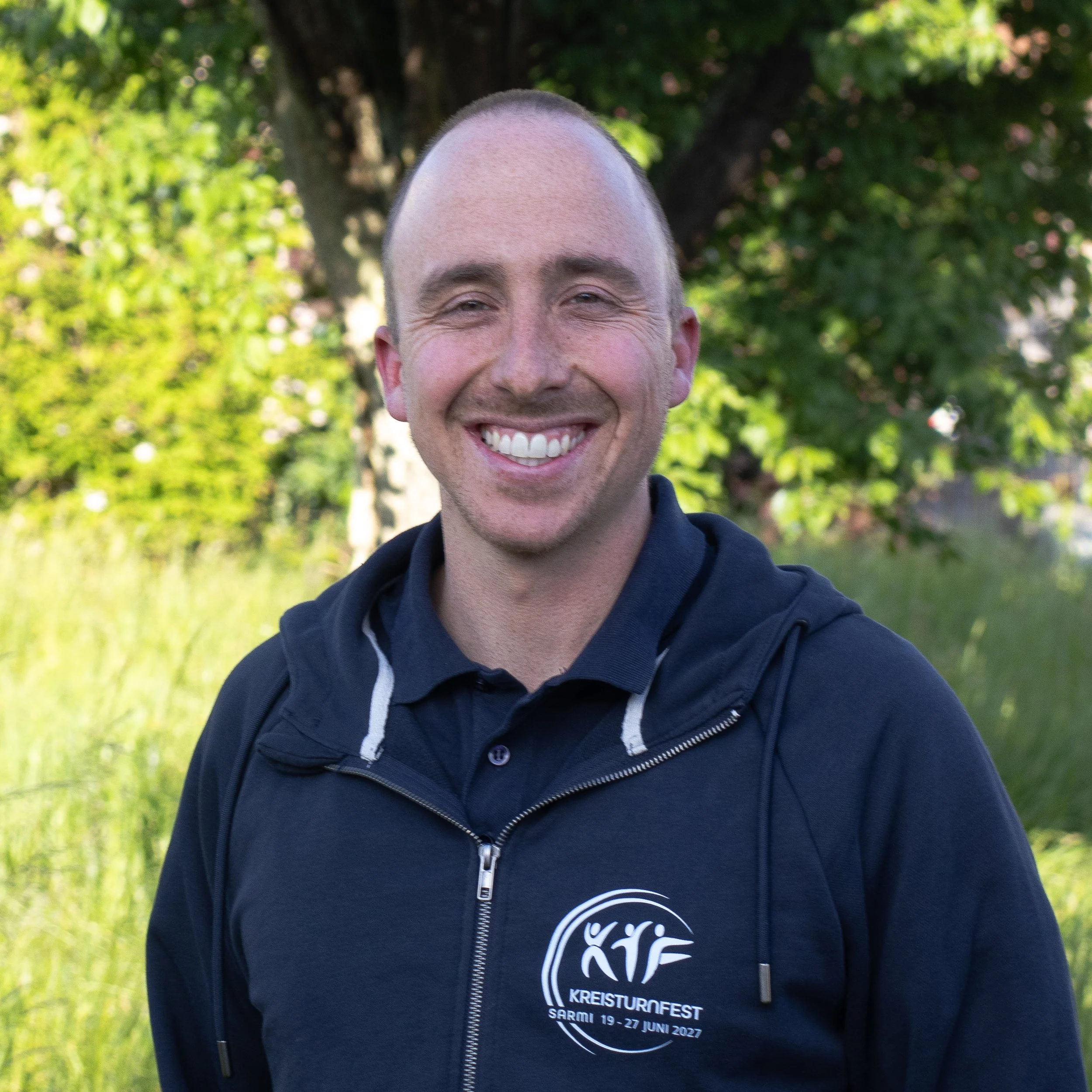 Ein fröhlicher Mann mit kurzem Haar, breitem Lächeln und weißen Zähnen, trägt ein dunkelblaues Jacke mit einem Logo, das für den Kreisturnfest Sarmi 2027 vom 19. bis 27. Juni steht, vor einem Hintergrund aus grünen Blättern und Bäumen.
