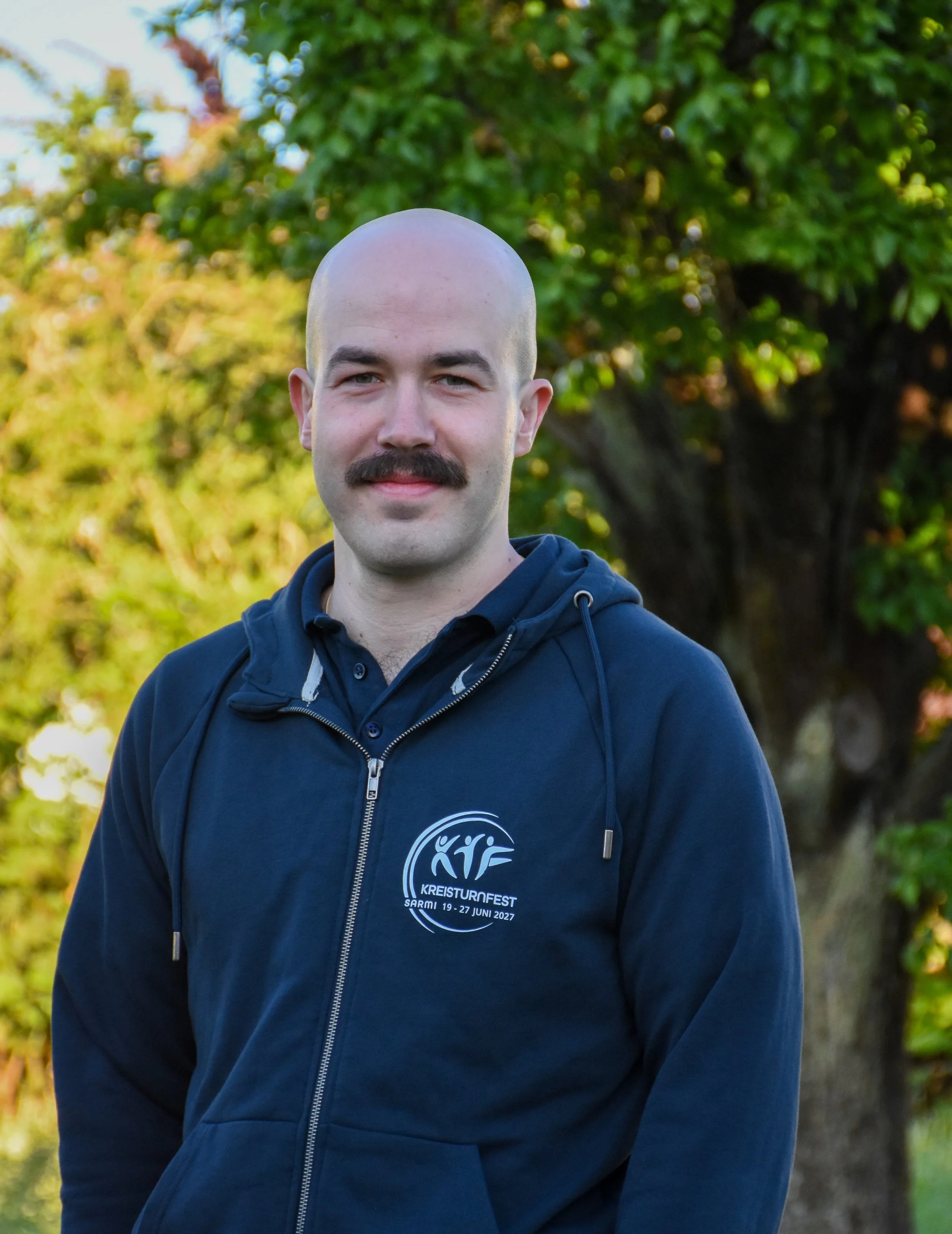 Ein Mann mit Bart und Glatze trägt einen dunkelblauen Hoodie mit einem Logo, das auf das Kreisturnfest 2021 vom 19. bis 27. Juni hinweist. Er steht vor einem Baum im Freien, umgeben von grünen Blättern, bei Sonnenlicht.