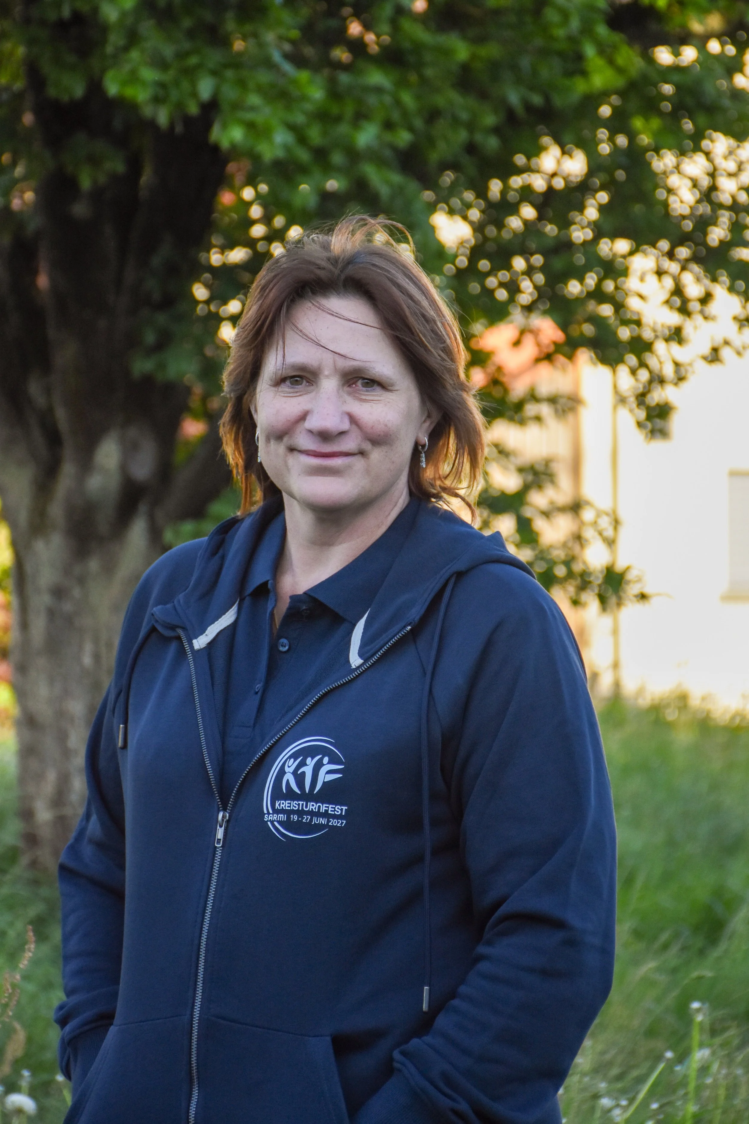 Ein Porträt einer Frau in einem blauen Hoodie, stehend im Freien vor einem Baum, bei Sonnenuntergang. Das Hoodie hat ein Logo und Text, der auf ein Kreisturnfest im Juni 2022 hinweist.