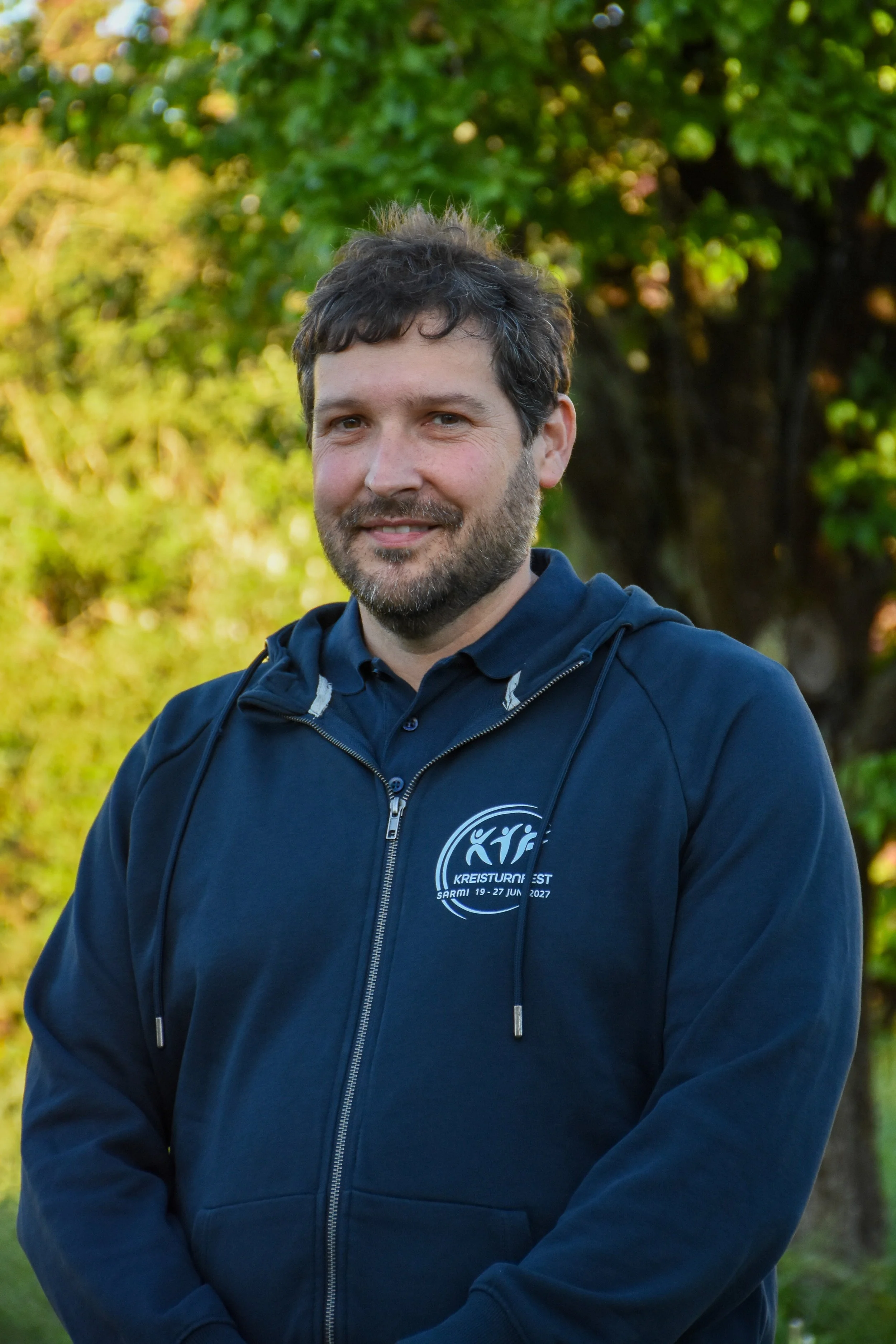 Ein Mann trägt eine dunkelblaue Jacke mit einem Logo, steht im Freien vor Bäumen mit herbstlichen Blättern, lächelt leicht.