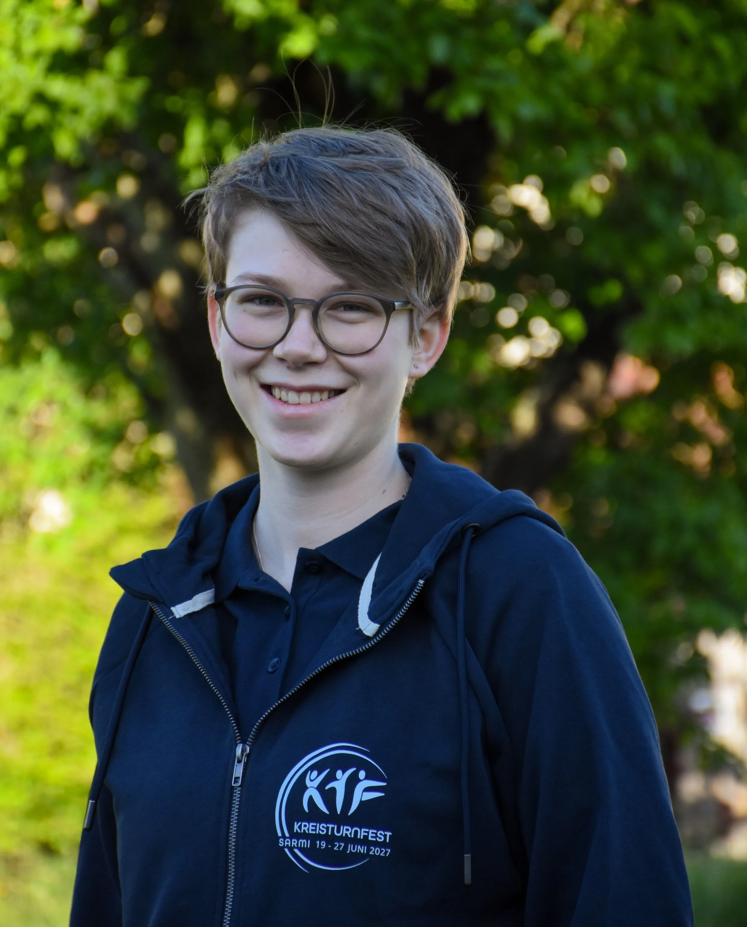 Junge Person trägt eine dunkle Hoodie mit Logo, im Hintergrund Bäume, lächelnd, kurze braune Haare, Brille, bei Tageslicht.