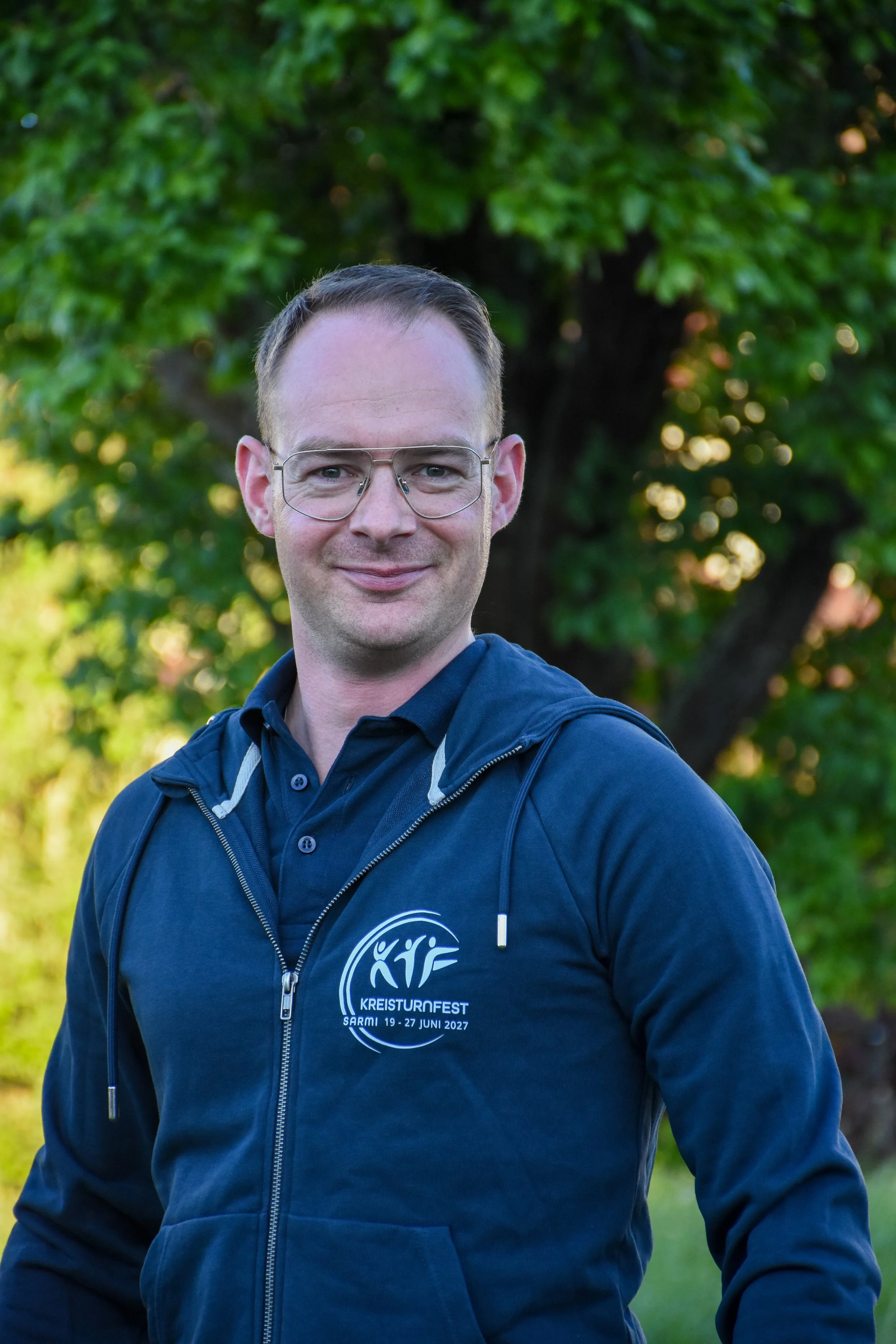 Ein Mann trägt eine blaue Jacke mit einem Logo, das den Text 'KREISTURNFEST SARMI 19-27 JUNI 2027' enthält, und steht vor einem grünen Baum im Freien.
